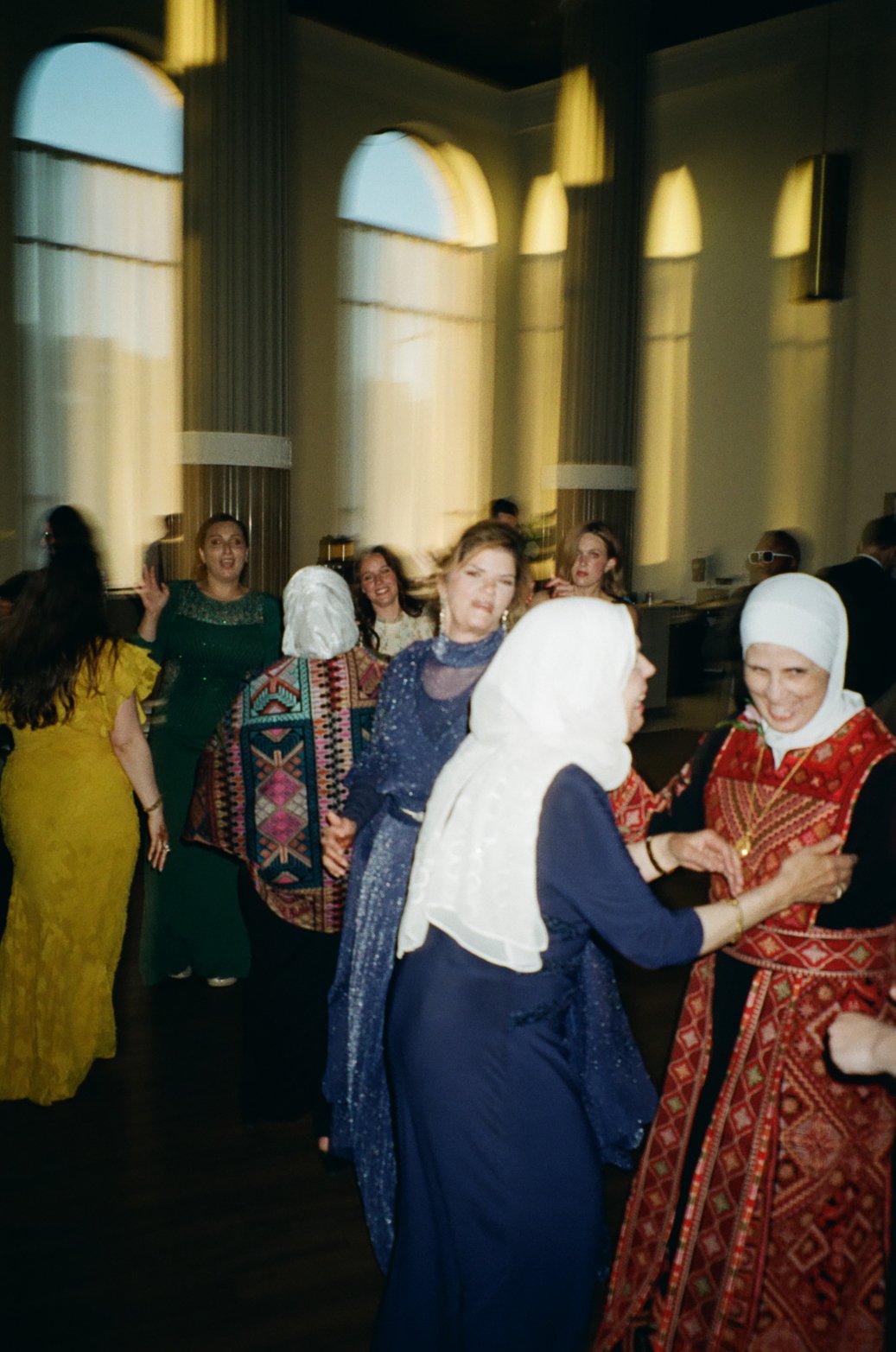 A group of women in colorful traditional and modern dresses dancing and chatting at an indoor celebration.