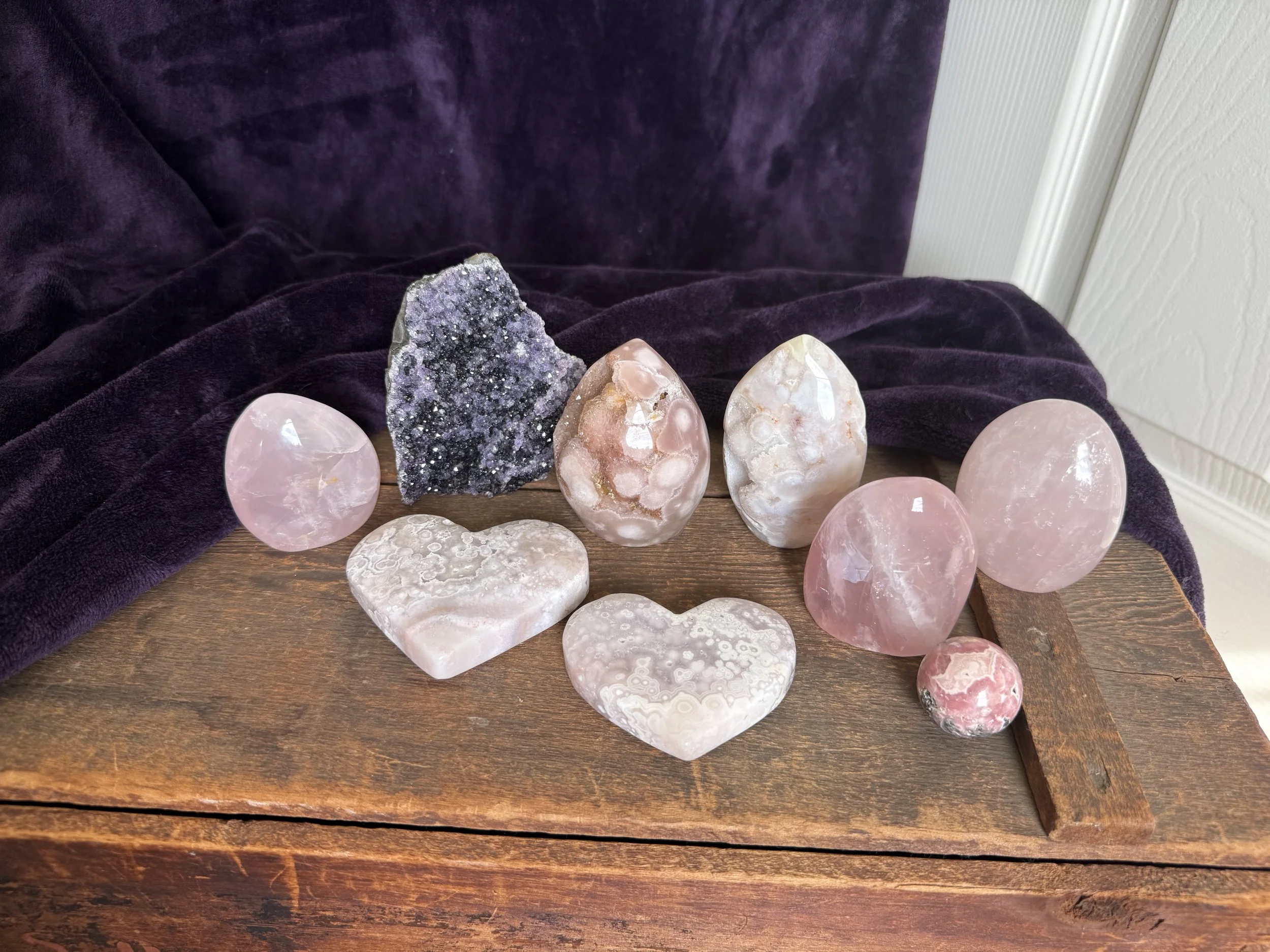 Various polished pink and purple crystals, including a dark druzy stone, arranged on a wooden surface with a purple cloth and white wall in the background.