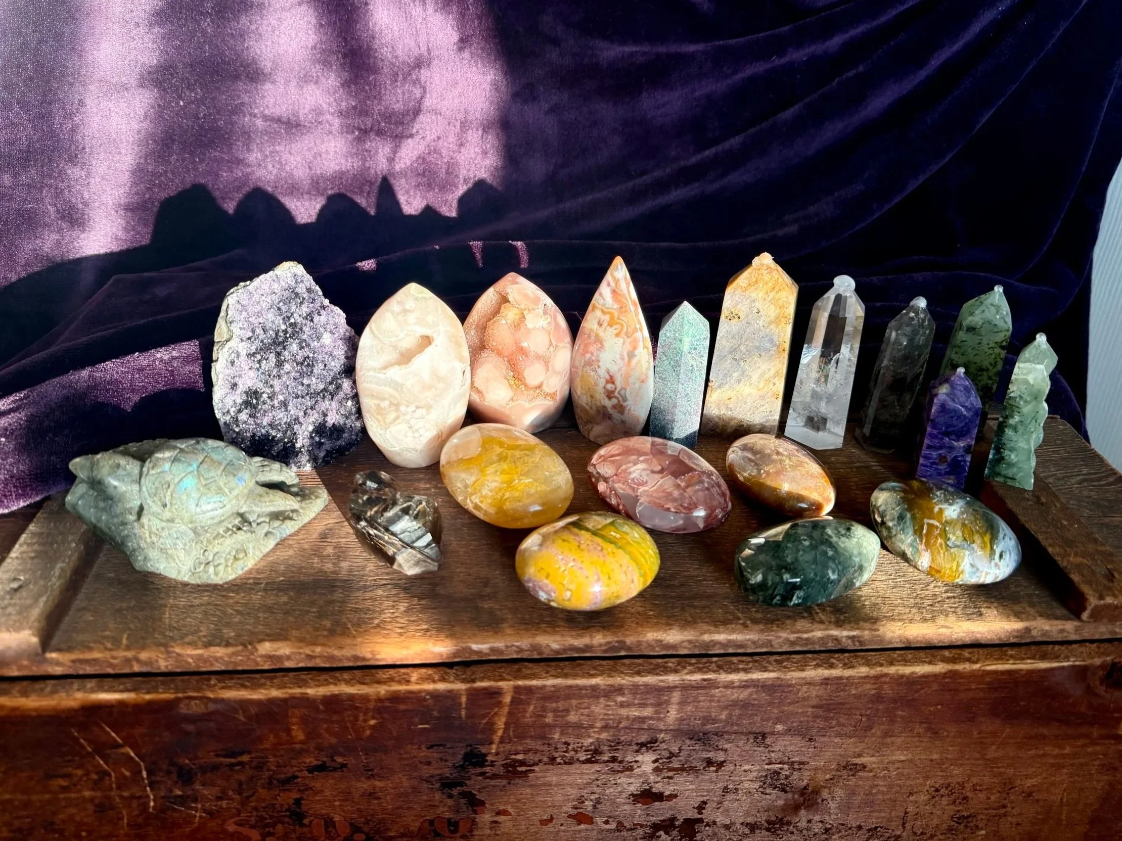 A display of various polished and raw gemstones and crystals on a wooden surface with a purple velvet fabric backdrop.