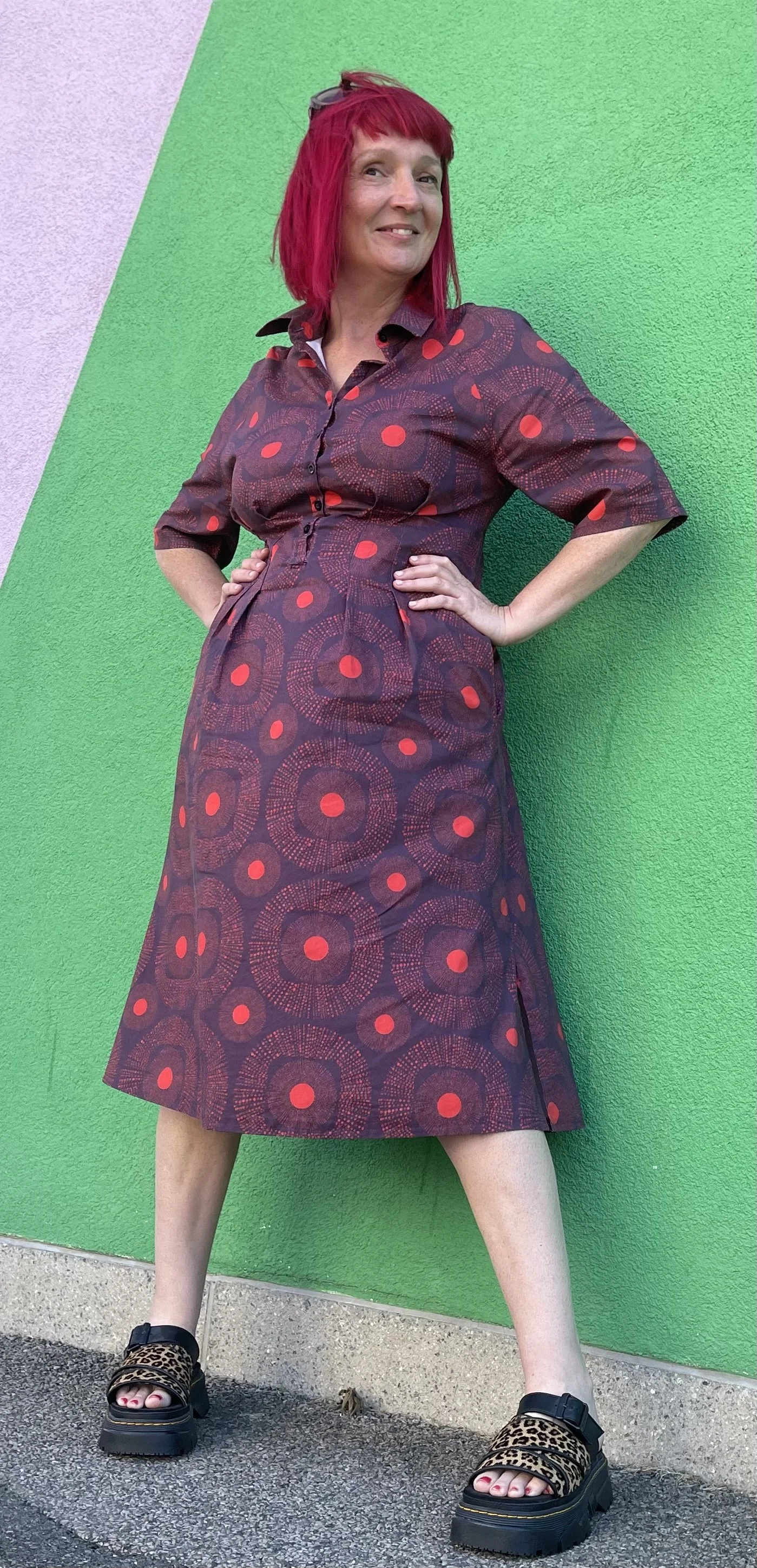 Woman with bright red hair wearing a dress she made herself using fabric she designed, leopard print platform sandals standing against a green textured wall with a pink section.