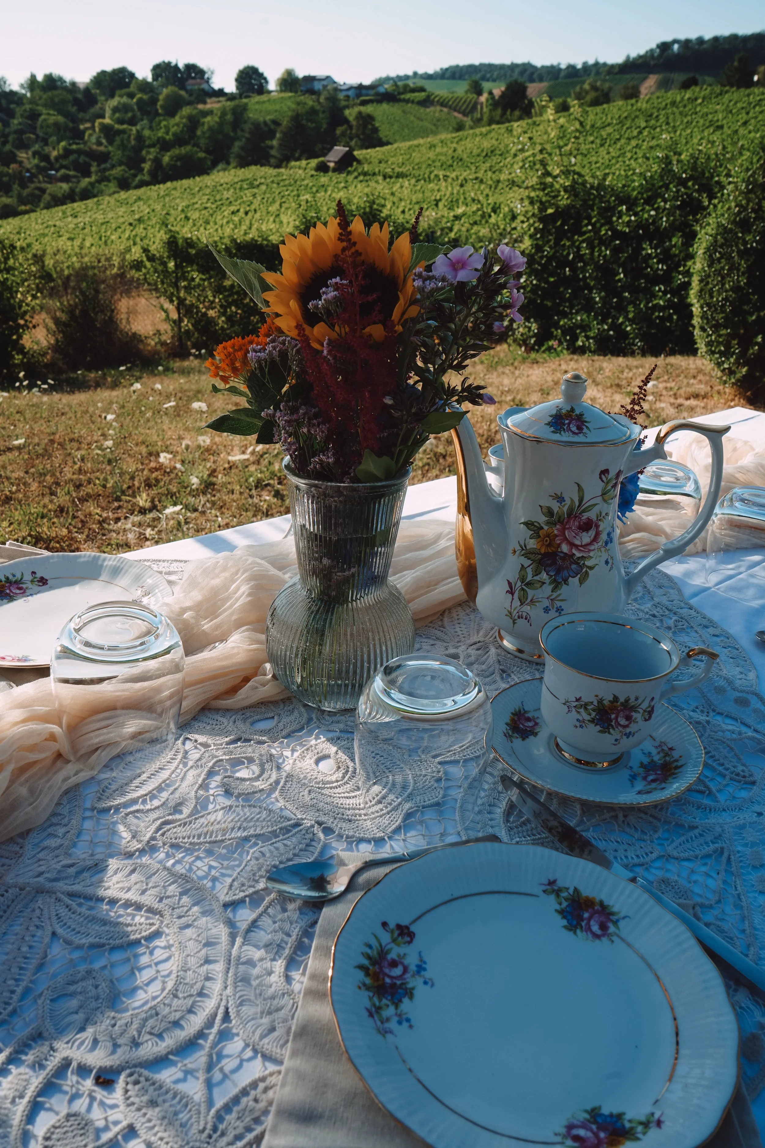 Frühstückstisch im Freien mit einer Vase voller bunter Blumen, einer eleganten Teekanne, Teetassen, Untertassen und Gläsern auf einem hellen, gehäkelten Tischdeckchen, mit landlichem grünen Weinberg und Hügel im Hintergrund bei sonnigem Wetter.
