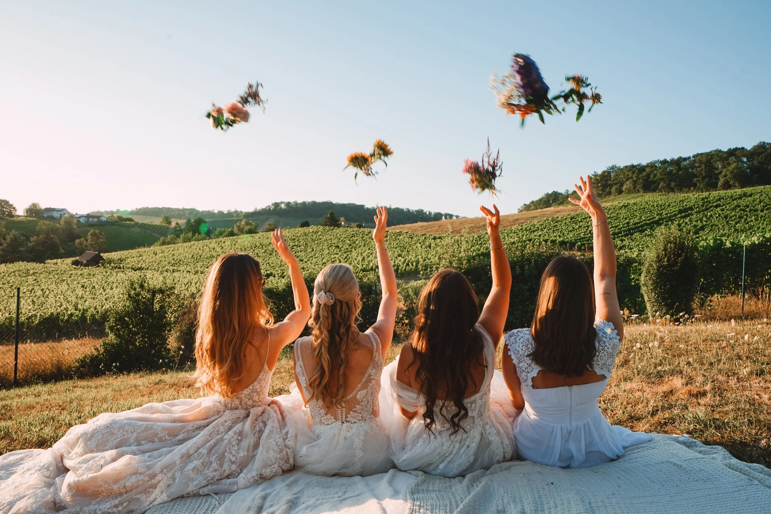 Sieben Frauen in weißen Kleidern sitzen auf einem weißen Tuch im Freien und werfen Blumen in die Luft, umgeben von einem landwirtschaftlichen Landschaft mit Hügeln und Weinbergen bei Sonnenuntergang.