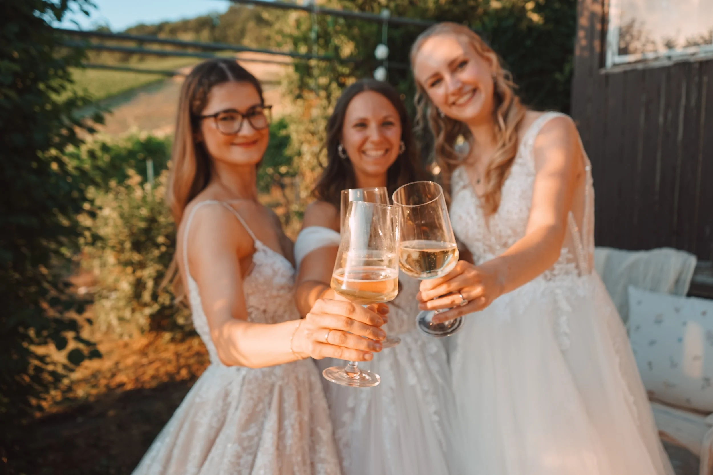 Drei Frauen in Hochzeitkleidern stoßen mit Sektgläsern an. Das Bild wurde draußen bei Sonnenuntergang aufgenommen.