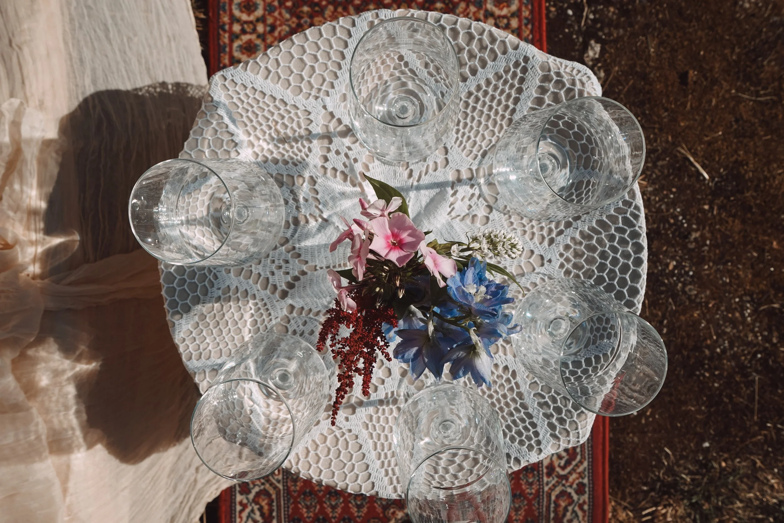 Leere Weingläser stehen auf einer Tischdecke mit Blumen. Der Tisch ist vor einem roten Teppich auf Erdboden. Schatten und Licht reflektieren auf den Gläsern.