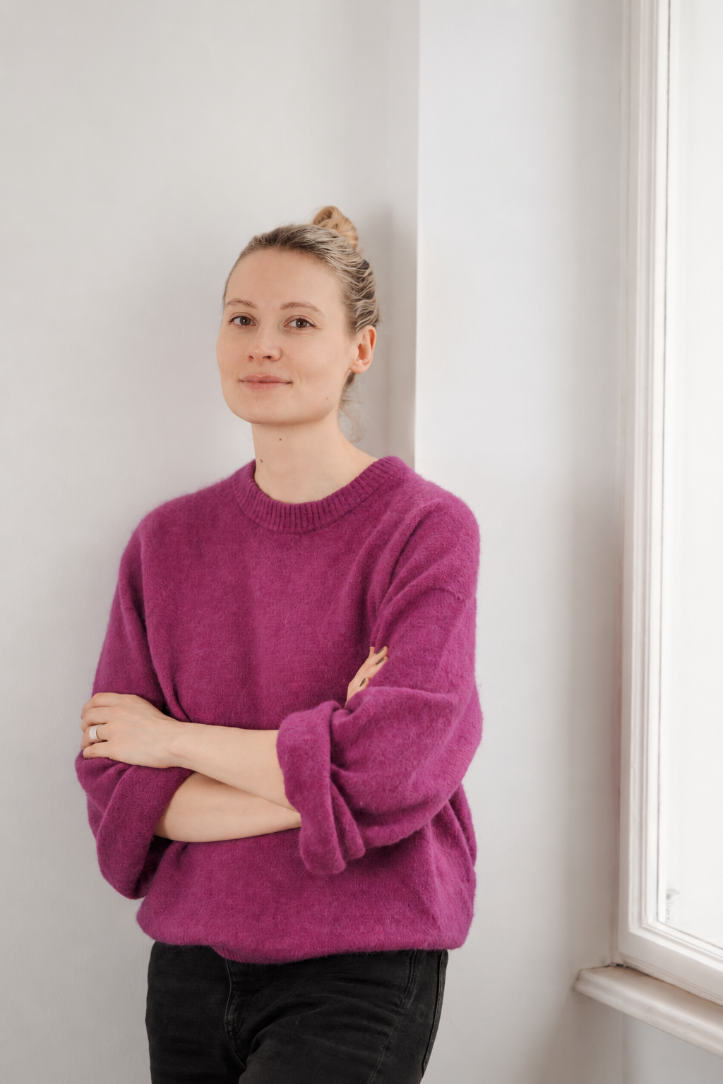 Eine junge Frau in einem magentafarbenen Pullover steht lächelnd mit verschränkten Armen vor einer weißen Wand neben einem Fenster.