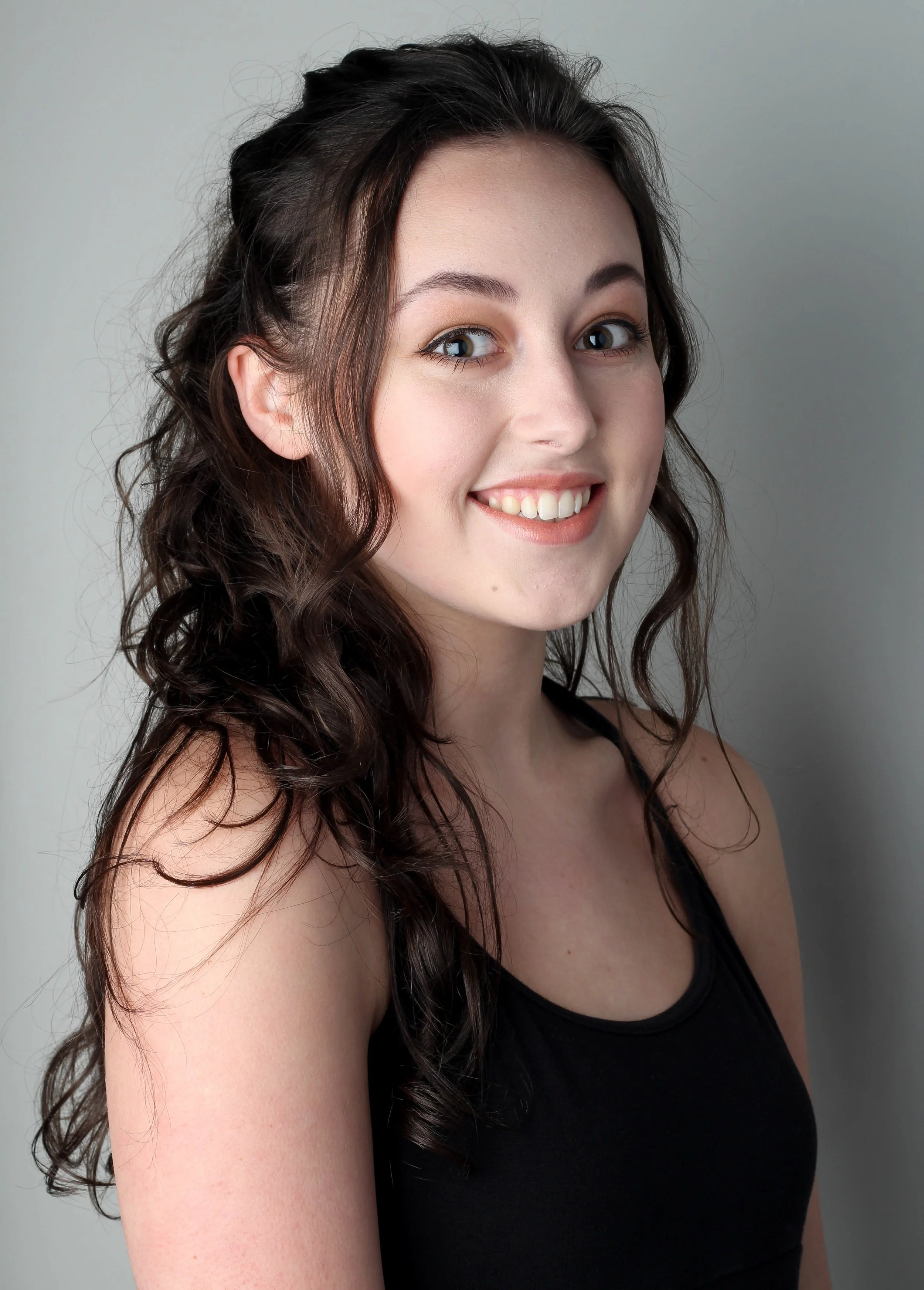 Portret van een lachende jonge vrouw met lang, golvend bruin haar en lichte huid, gekleed in een zwart top, tegen een effen lichte achtergrond.