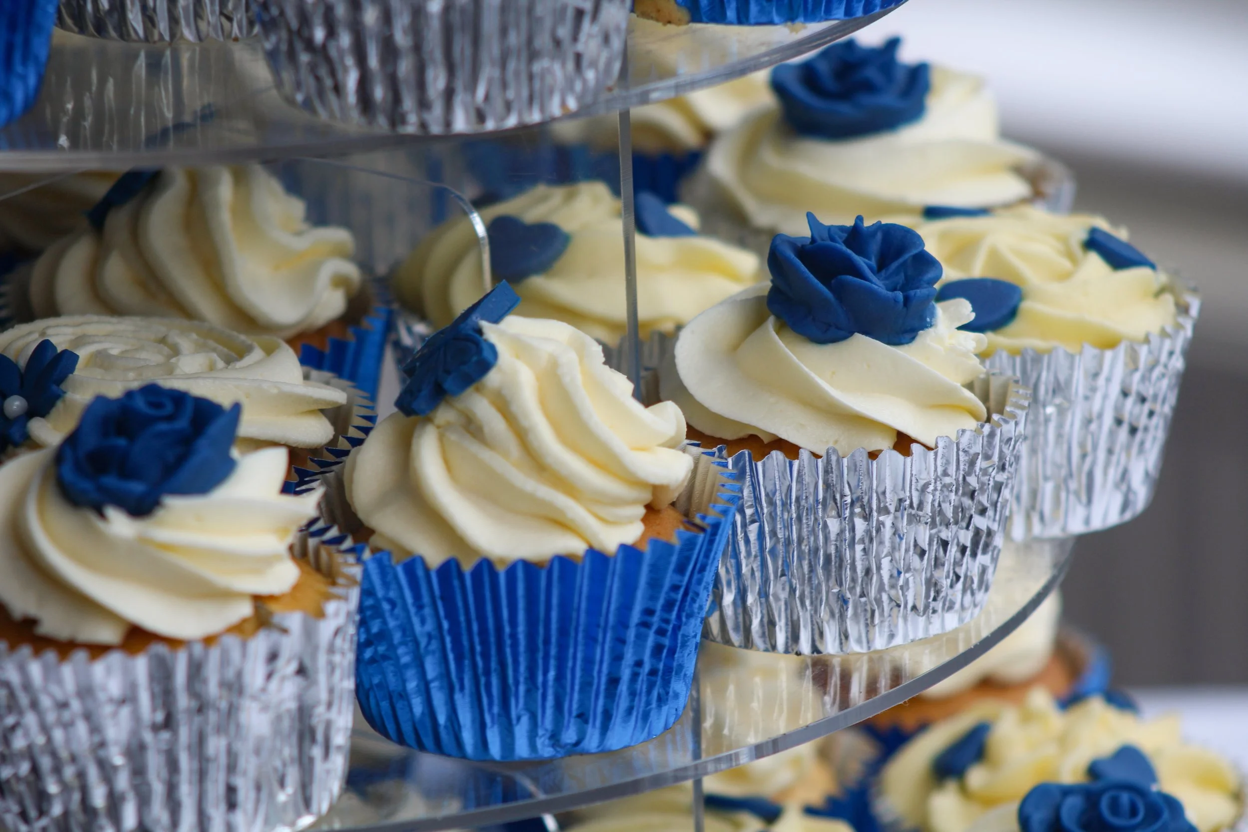 Cupcakes met witte frosting en blauwe rozenversiering in blauwe en zilveren papiertjes, op een glazen etagère