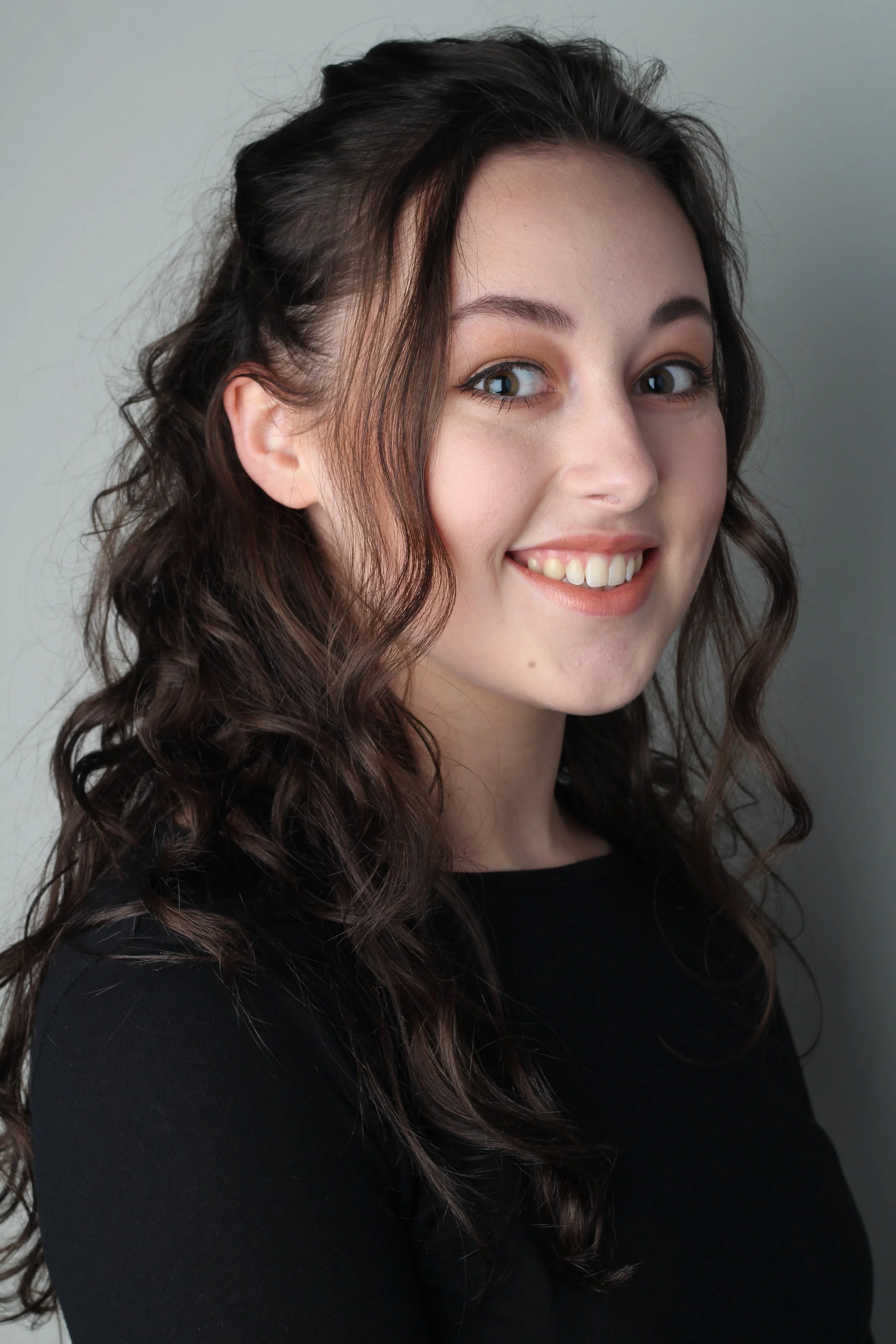 Portret van een vrouw met lang, bruin, gekruld haar, lachend met witte tanden, dragend zwarte top, staand voor een neutrale grijze achtergrond.