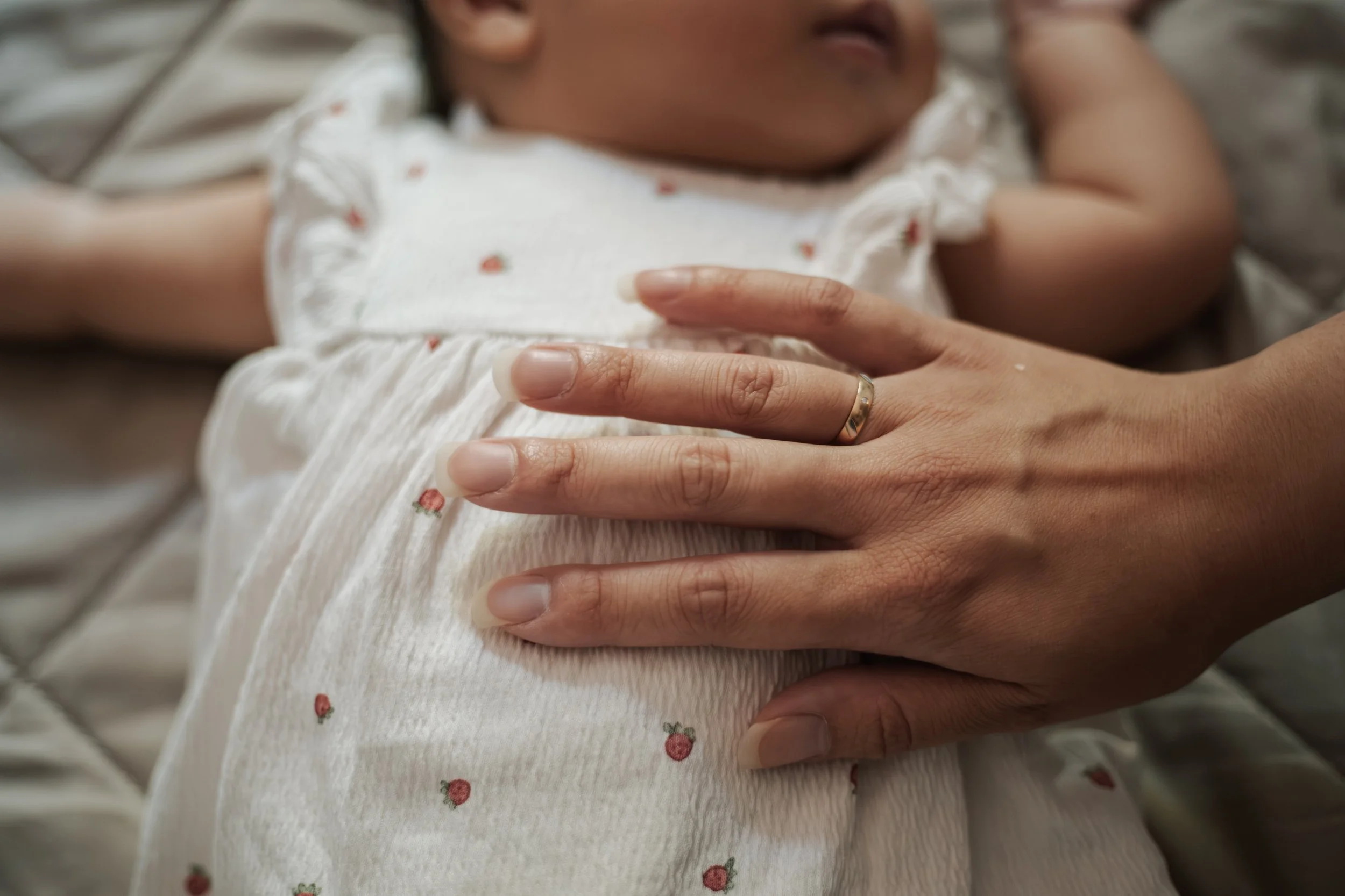 Close-up van een baby die op een volwassene ligt, met de hand van de volwassene op de buik van de baby. De baby draagt een wit rompertje met een kleine bloemenprint.