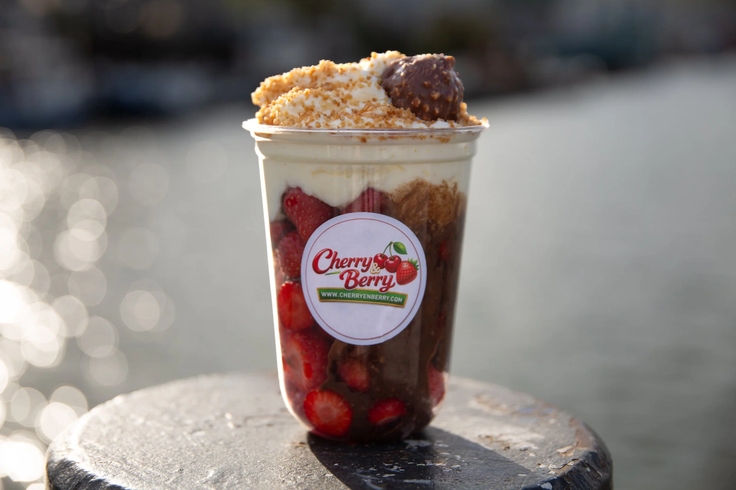 Een doorzichtig plastic bekertje gevuld met aardbeien, witte room, chocolade, en gebroken biscuit. Het bekertje heeft een sticker met de tekst 'Cherry & Berry' en een websiteadres. Op de achtergrond is een waterachtige omgeving met weerspiegelingen e