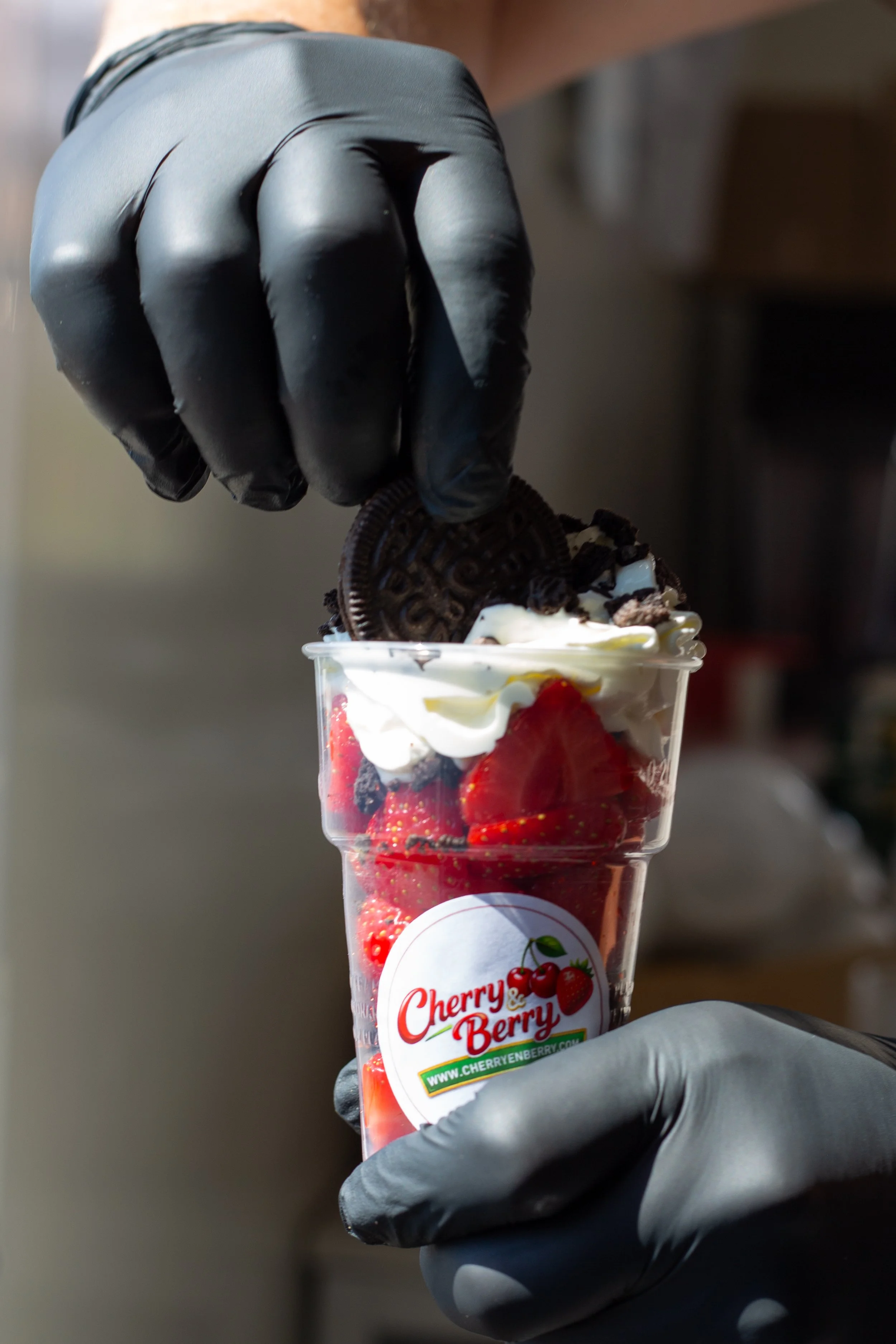 Persoon met zwarte handschoenen houdt een doorzichtige beker met ijs, aardbeien, slagroom en een Oreo-koekje boven een dessert.