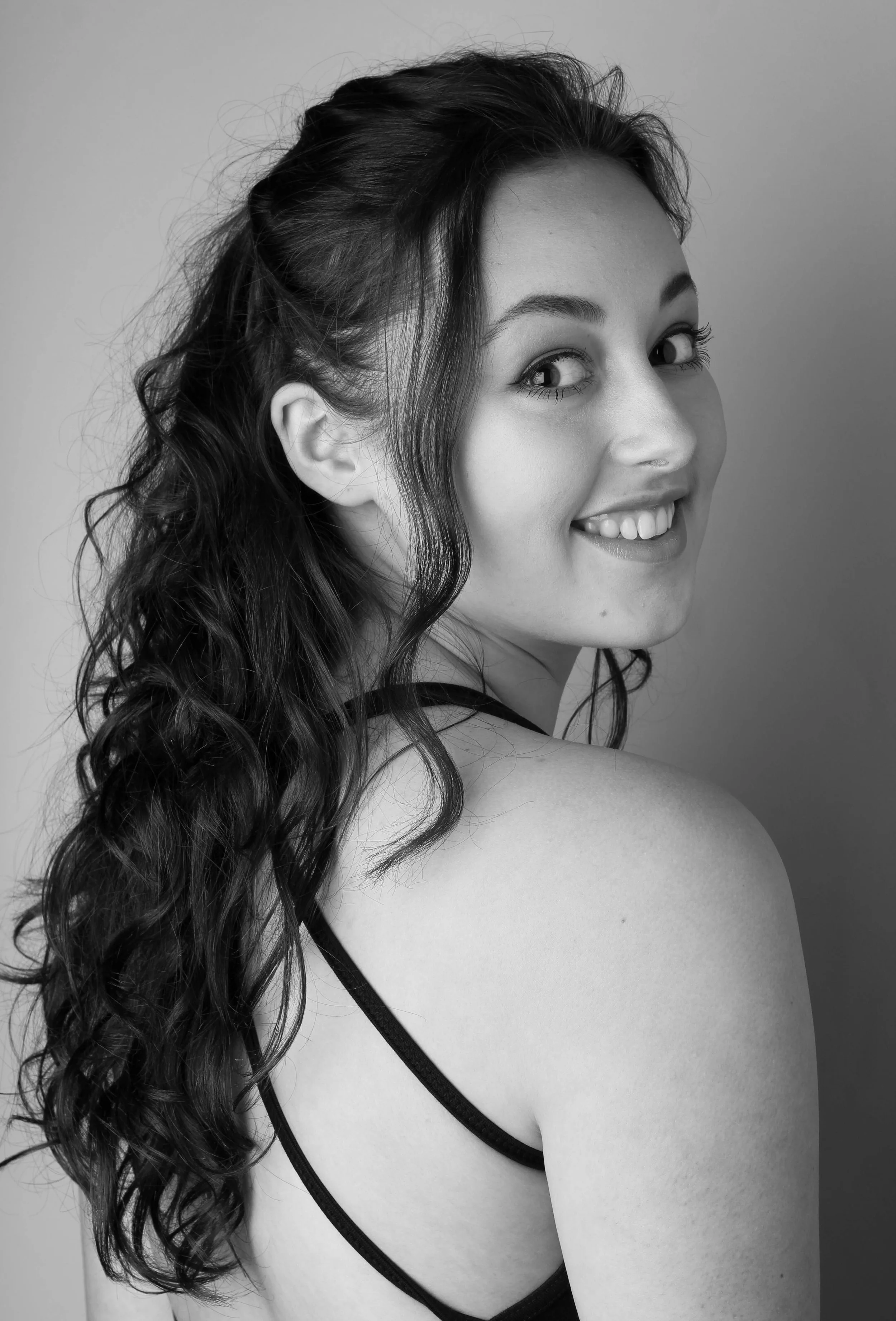 Zwart-wit foto van een vrouw die naar de camera glimlacht, met lang, golvend haar en een zwart spaghetti-strap top.