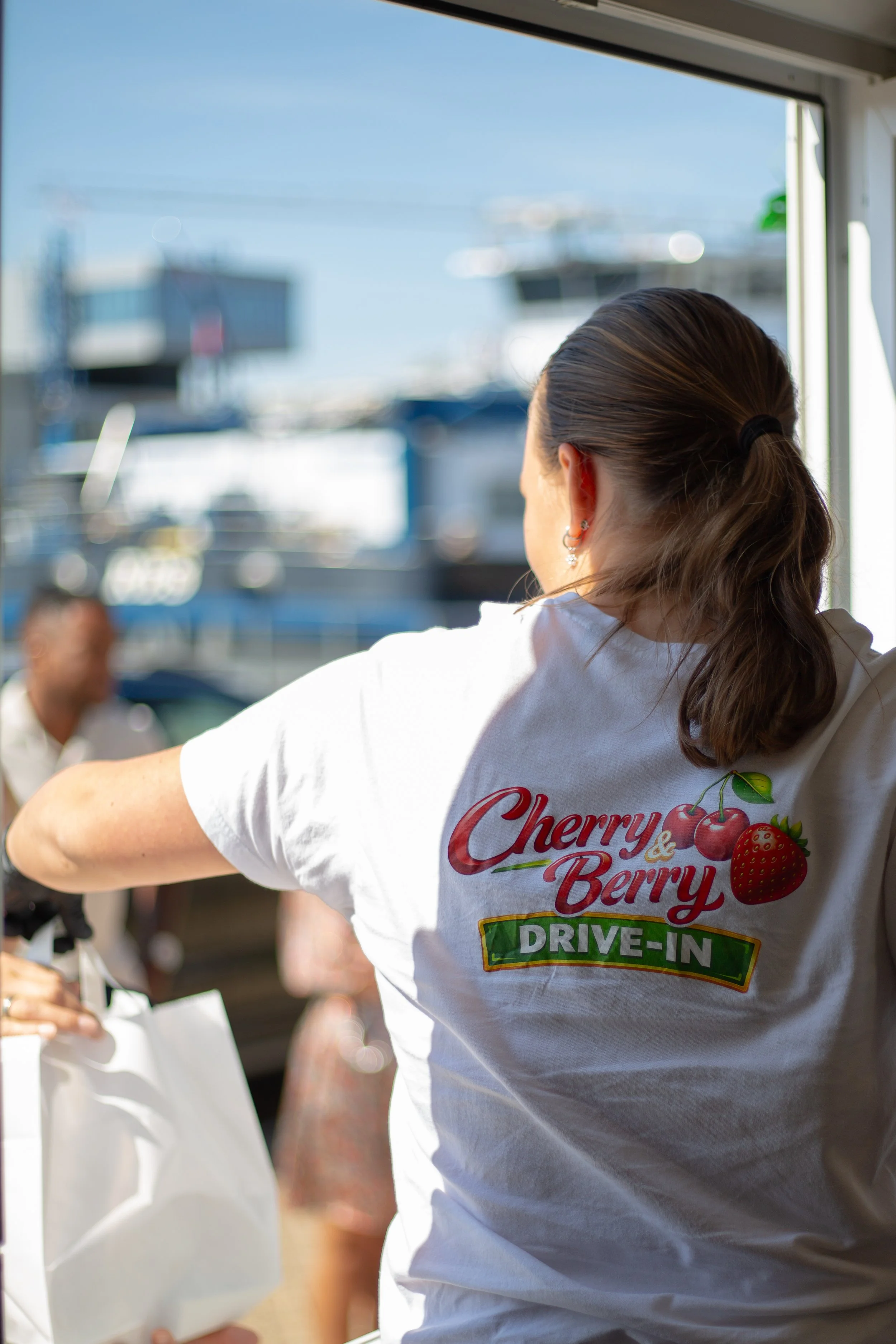 Een persoon ziet uit een raam met een shirt dat zegt 'Cherries & Berries' en 'Drive-In', terwijl anderen in de achtergrond bestellingen ontvangen.