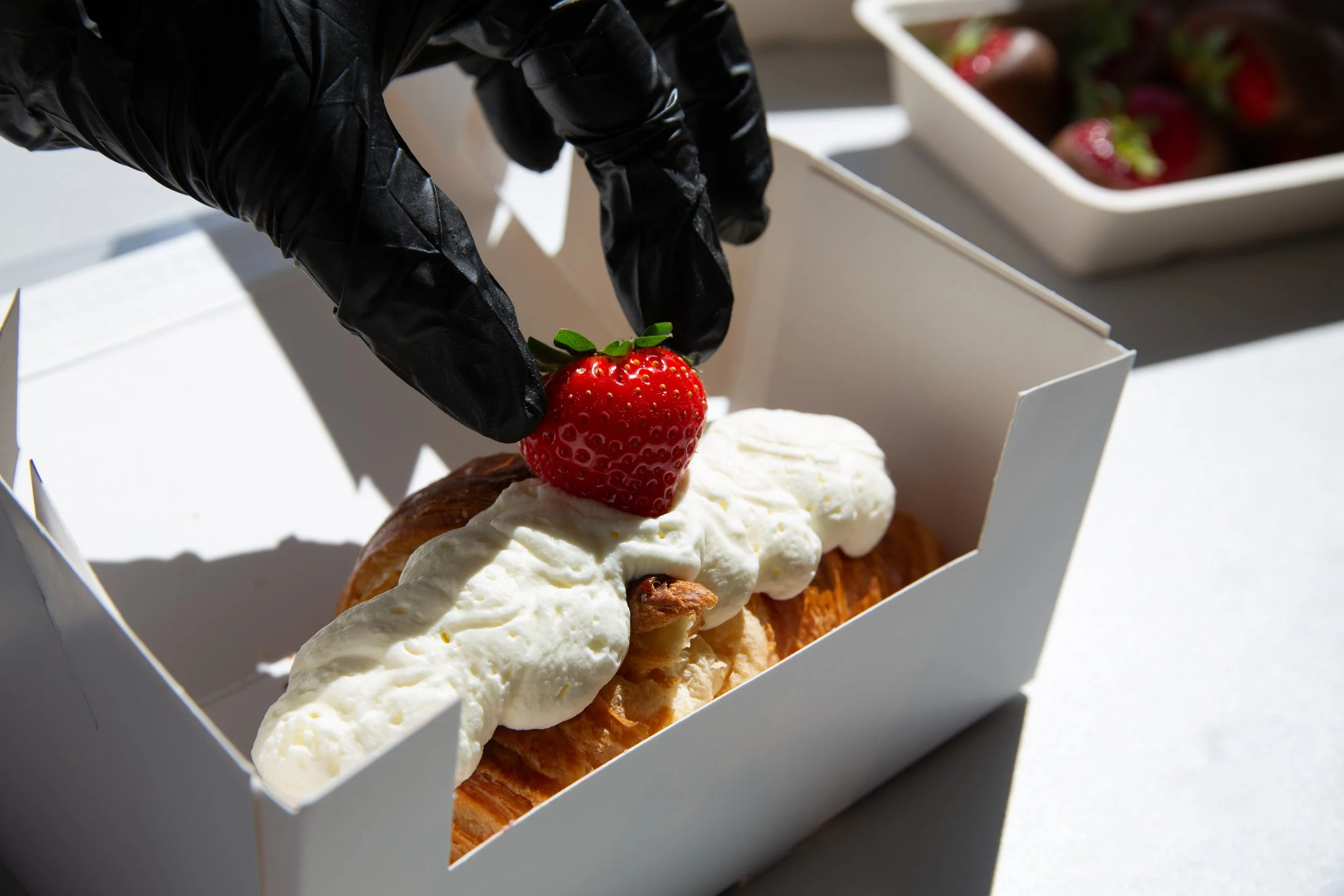 Close-up van een dessert met slagroom, een aardbei en stukjes noot in een witte doos. Iemand draagt zwarte handschoenen om het aardbeitje te plaatsen.
