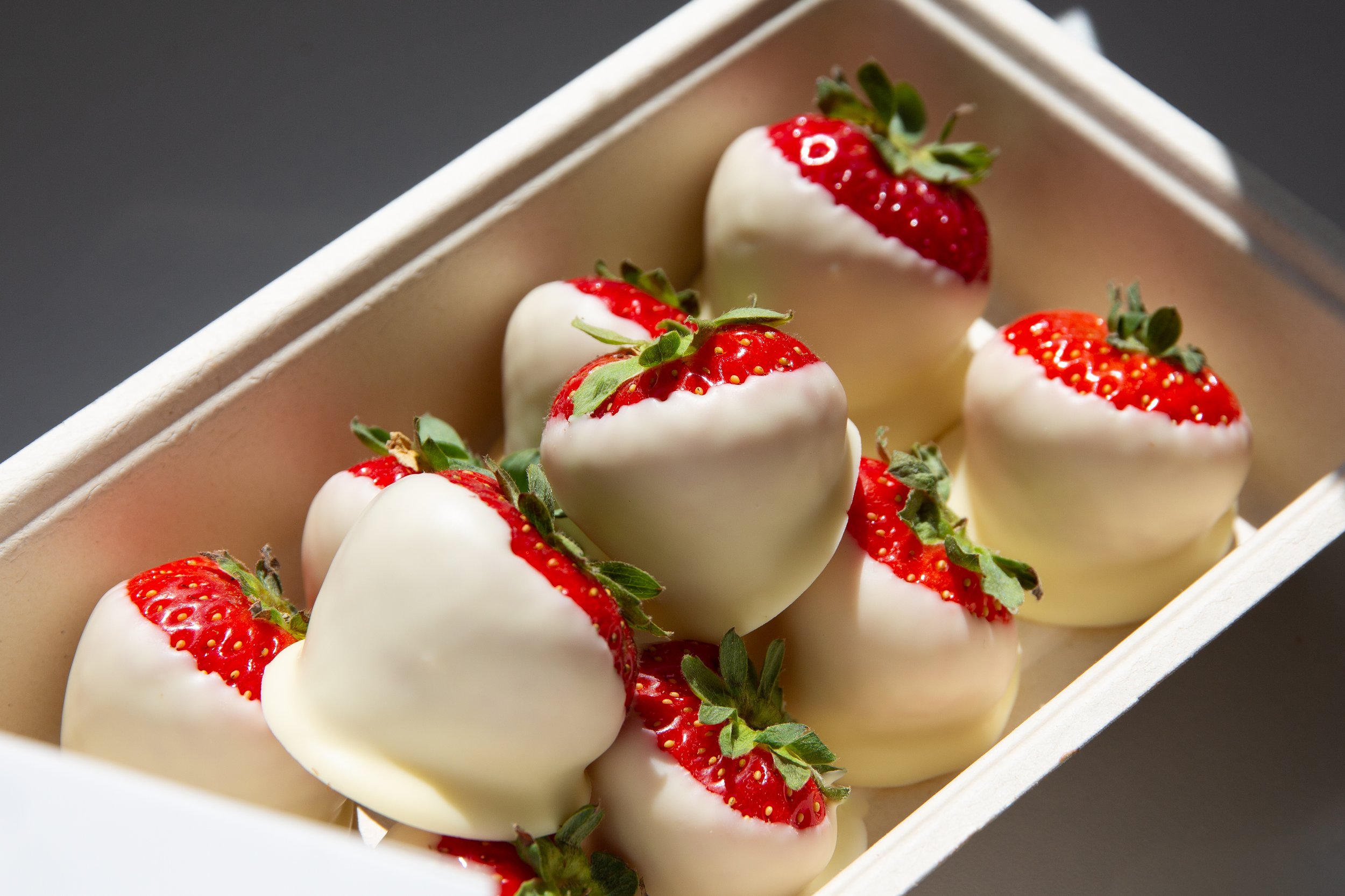A box of white chocolate-covered strawberries with red strawberries and green leaves inside.