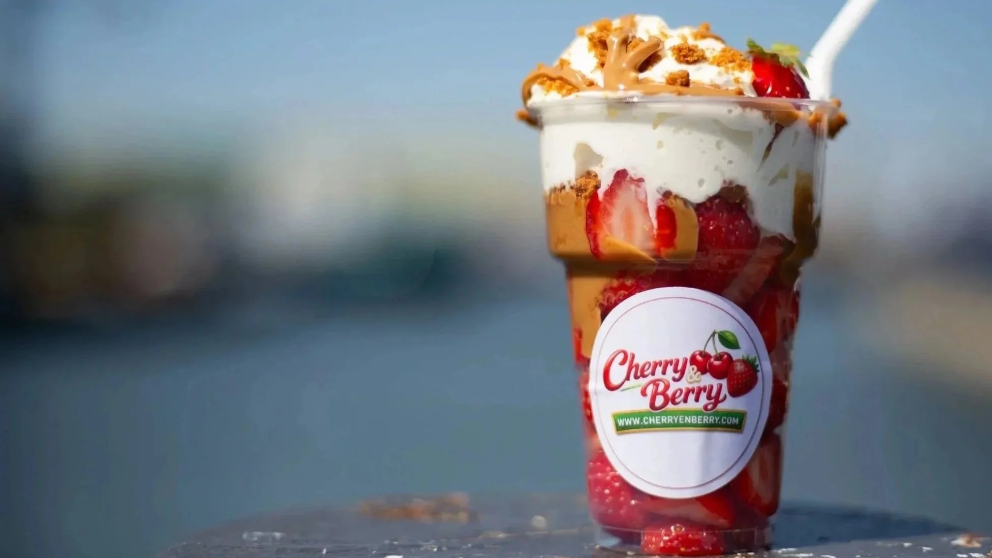 Een glas met een dessert met aardbeien, yoghurt, crumble en karamel, met een rietje en label van Cherry & Berry.