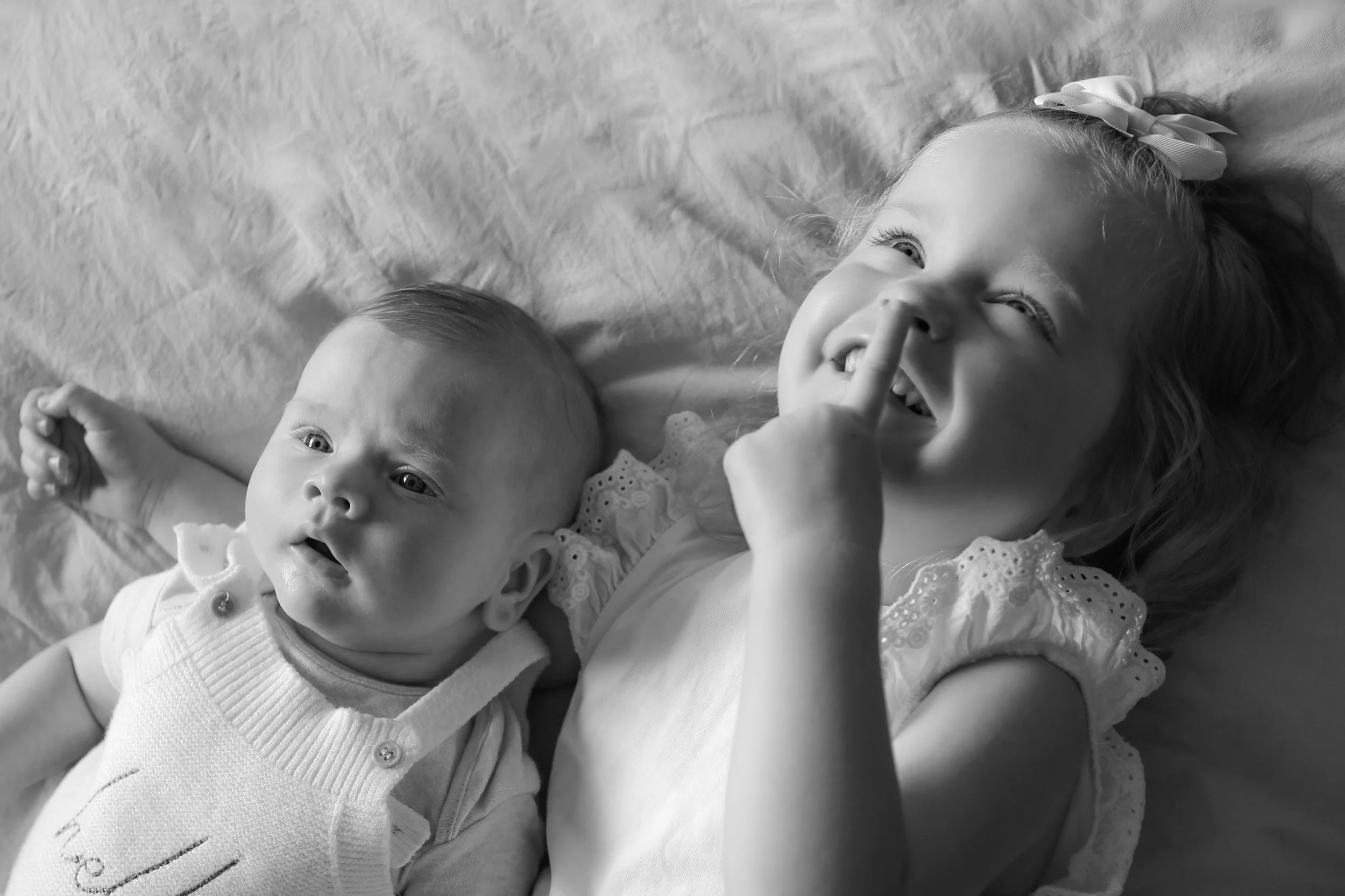 Lachende jonge meid ligt naast een baby op een zachte ondergrond, de meid wijst met haar vinger naar haar neus.