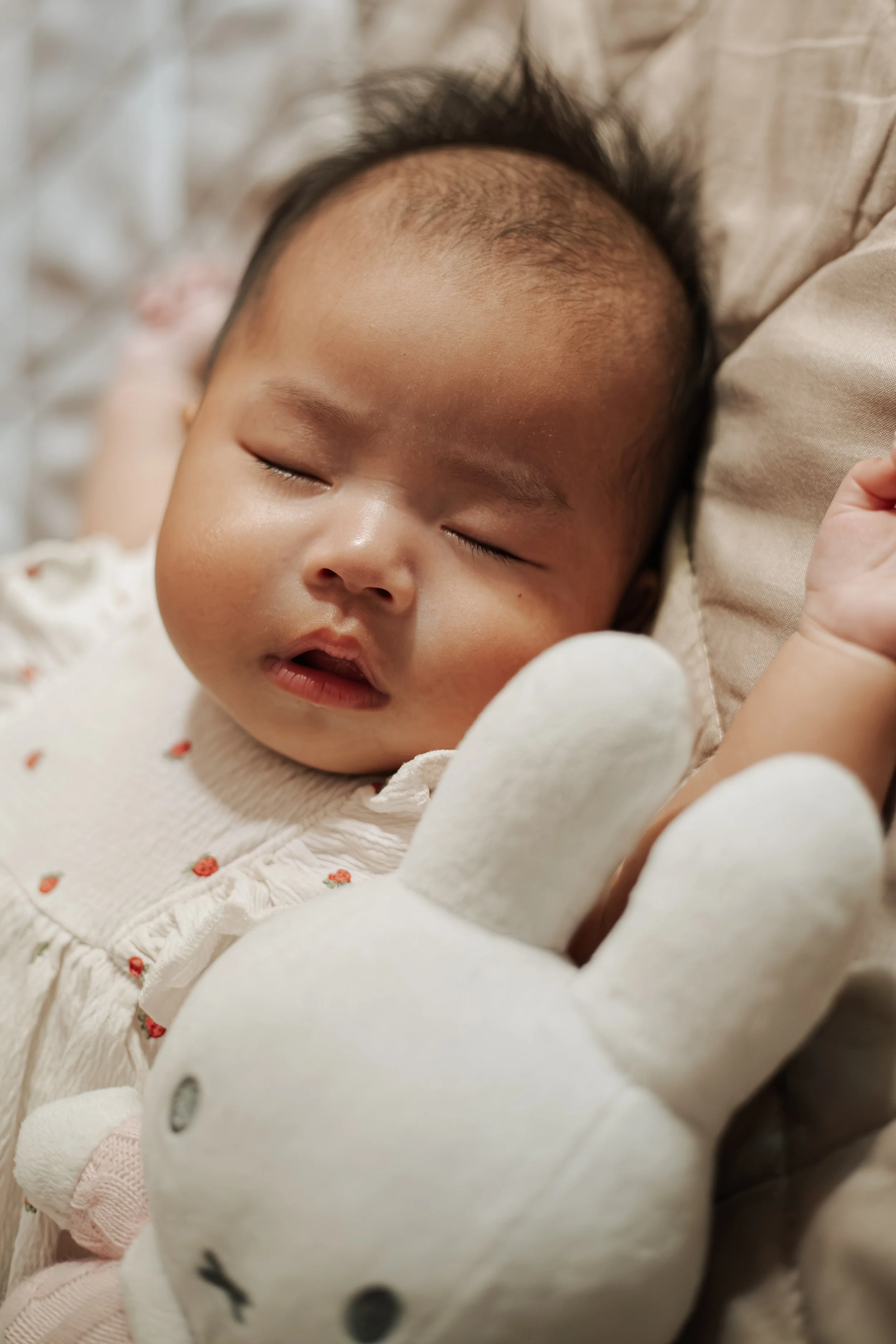 Een close-up foto van een slapende baby met donkere haartjes, dichtgeklemde ogen en een lichtbruine huid, liggend op een bed met zachte lakens.