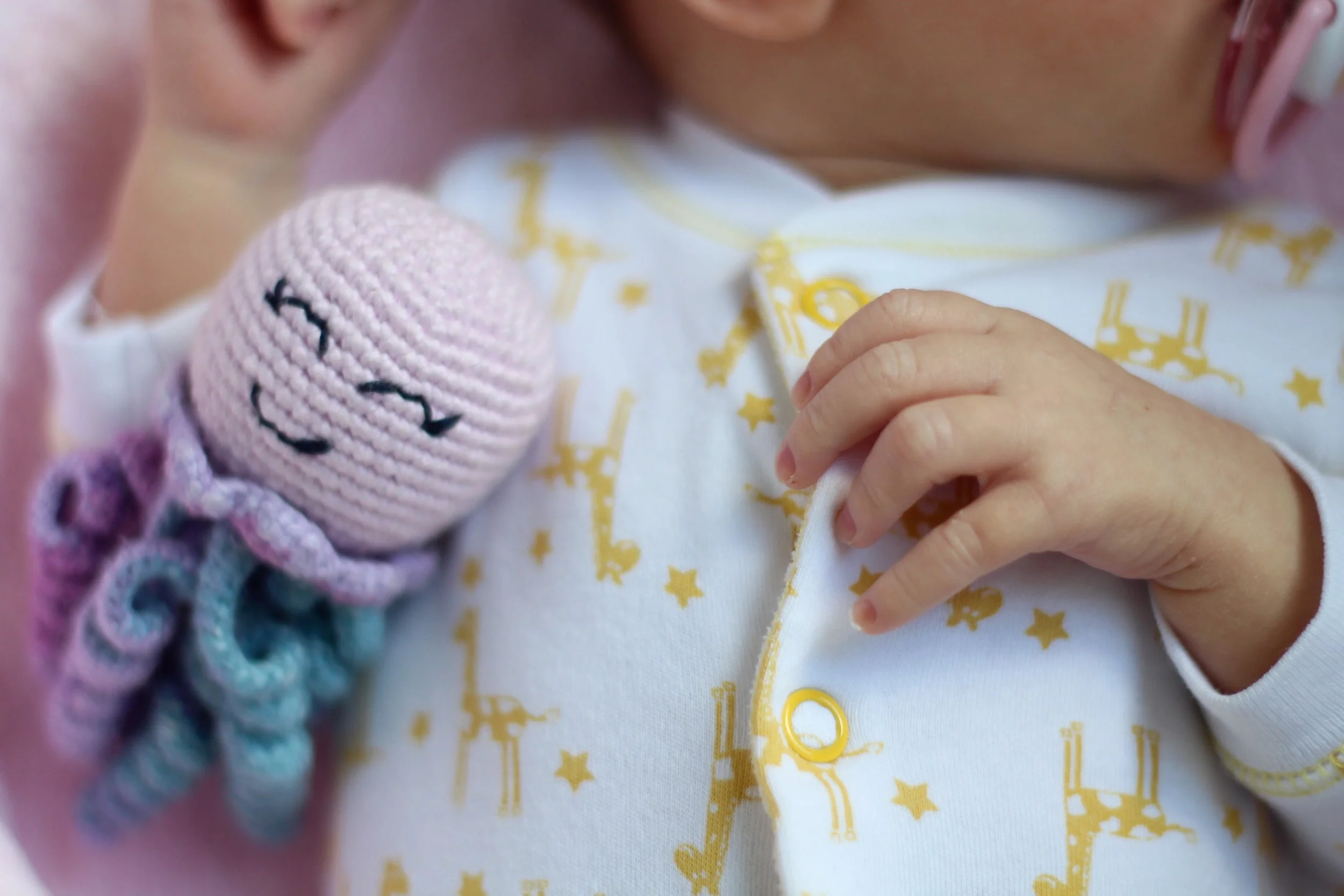 Close-up van een baby die rust op een babydekentje met gele sterren en astronauten, met een hand die zachtjes op de borst van de baby ligt, en een roze gebreide knuffel met de naam 'Kiki'.
