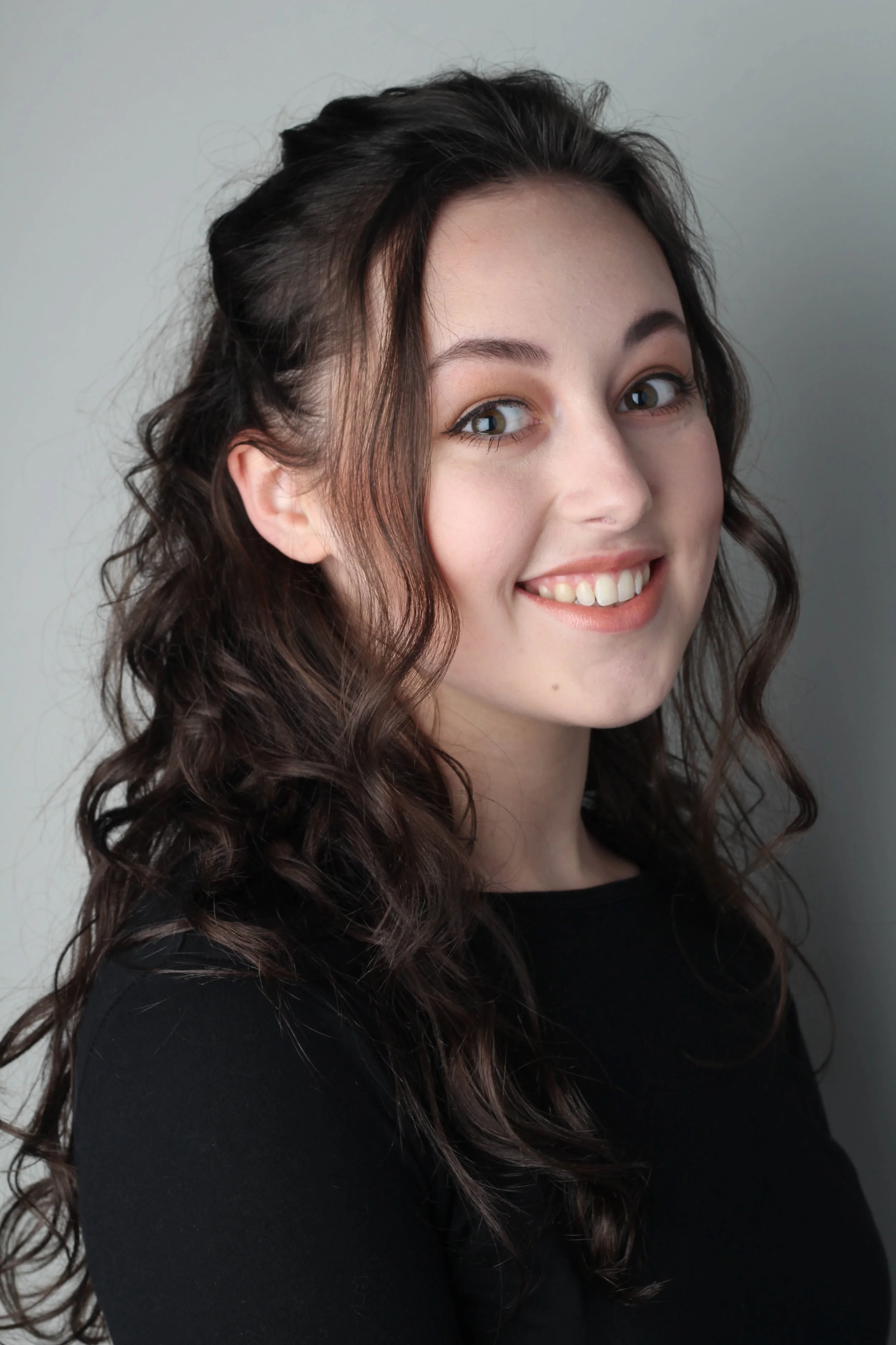 Portret van een jonge vrouw met lang, krullend bruin haar, lachend en kijkend naar de camera, gedragen in een zwarte top, tegen een lichte effen achtergrond.