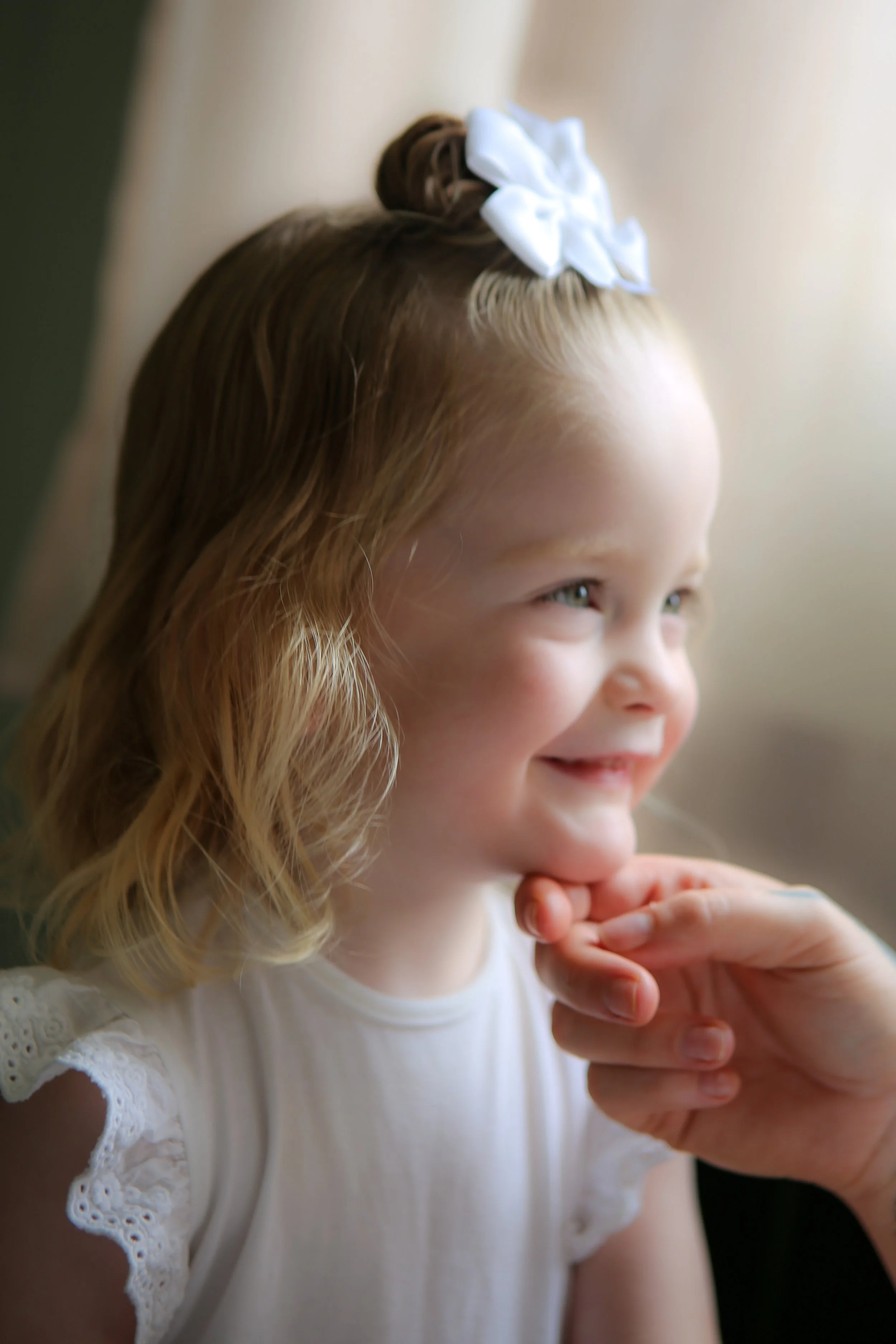 Close-up van een lachend meisje met blonde haar, witte kanten jurk, haar in een knot met een witte strik, en een hand die onder haar kin rust.