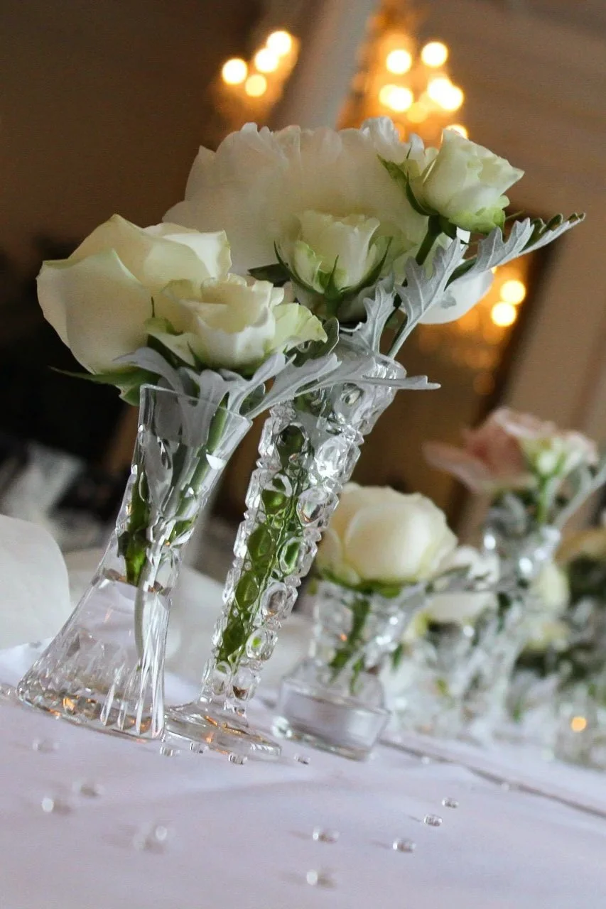 Witte bloemen in heldere kristallen vazen op een witte tafel met kleine glanzende decoraties, mogelijk voor een bruiloft of feest, met een warme, sfeervolle achtergrond met lichtjes.