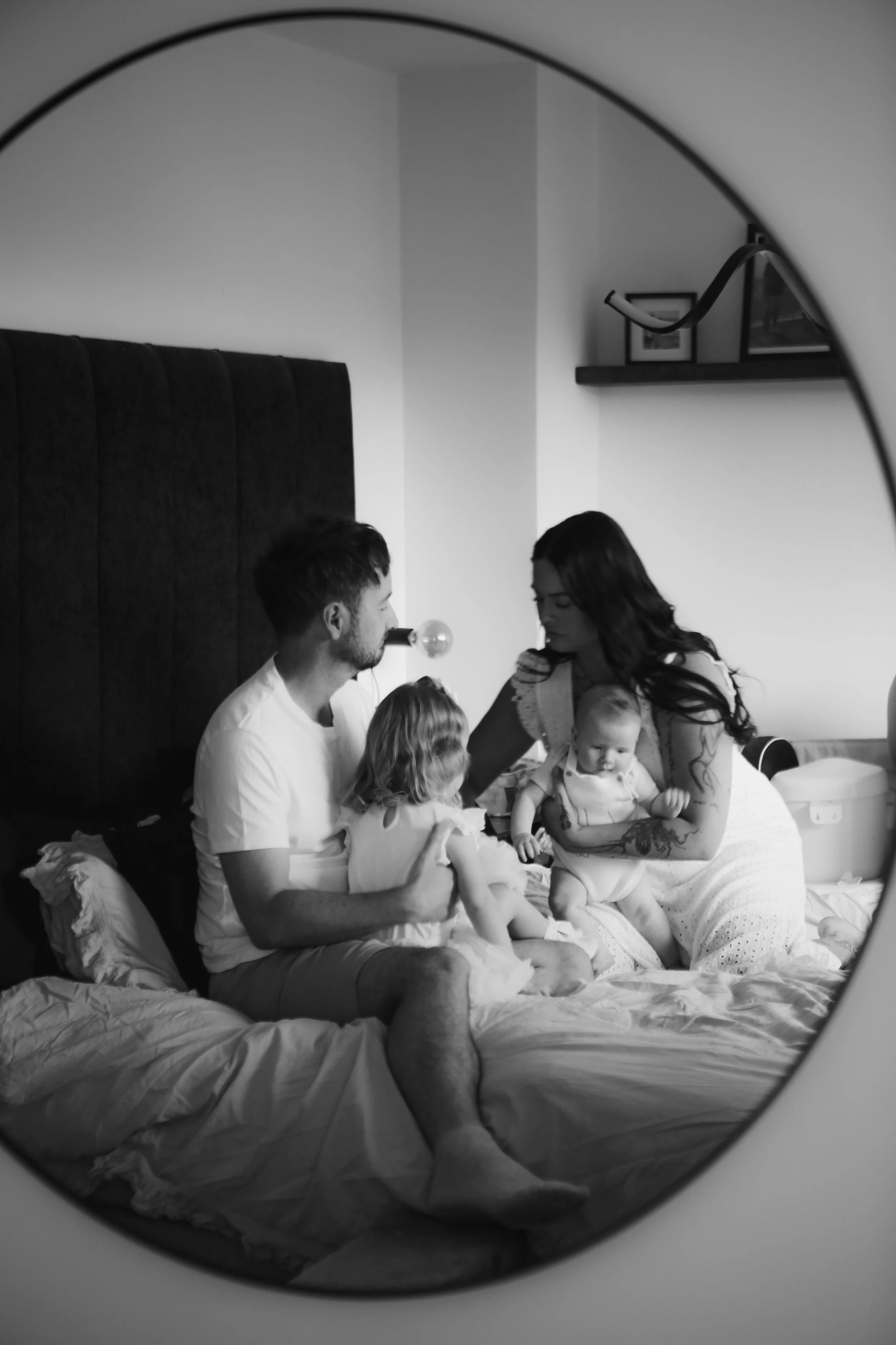 Een zwart-wit foto van een gezin in een slaapkamer, gezien via een ronde spiegel. Het gezin bestaat uit een man, een vrouw en twee jonge kinderen, die samen op het bed zitten en knuffelen.