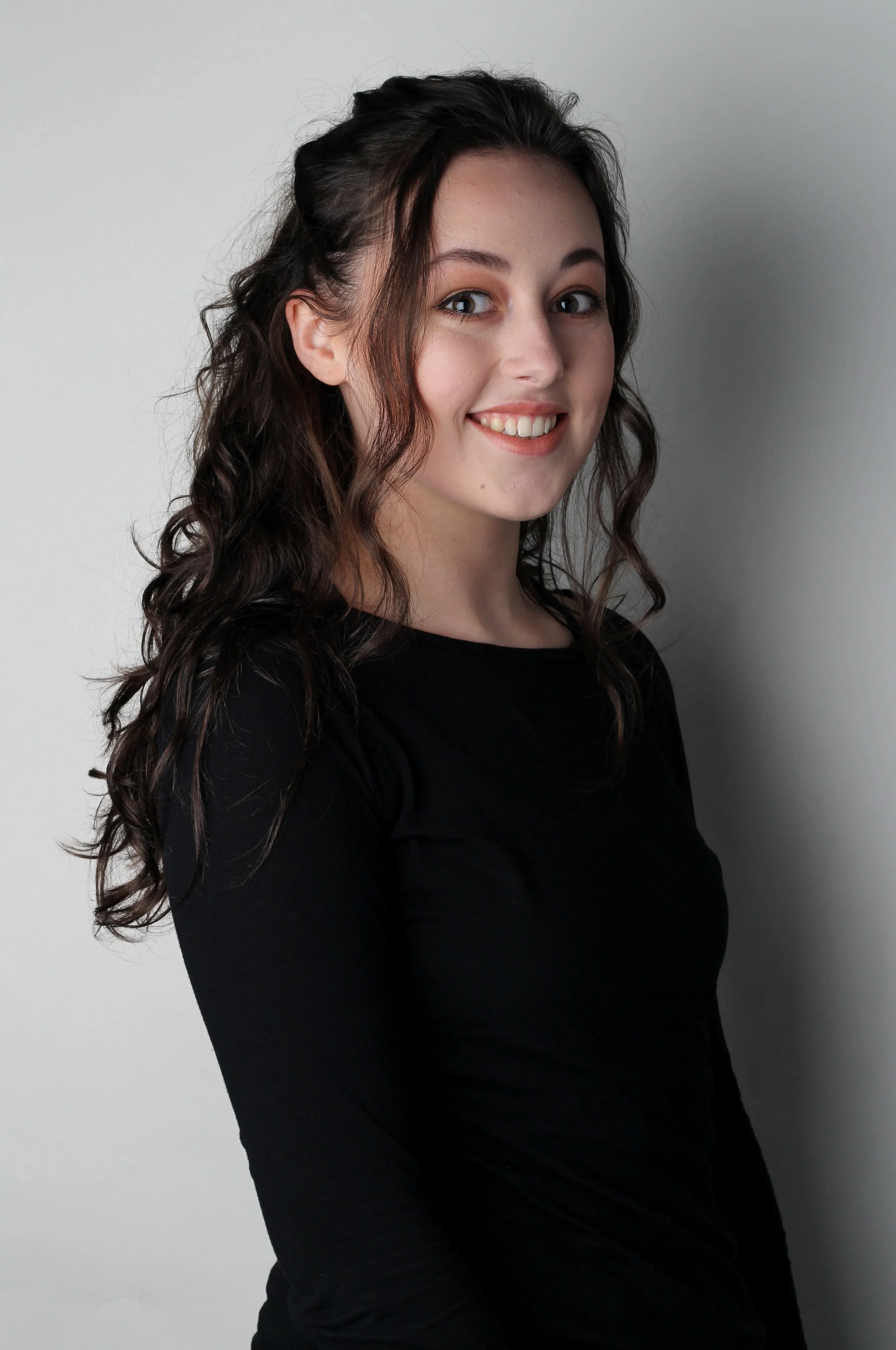 Portret van een jonge vrouw met lang, krullend bruin haar, glimlachend en gekleed in een zwarte top, staande voor een lichte achtergrond.