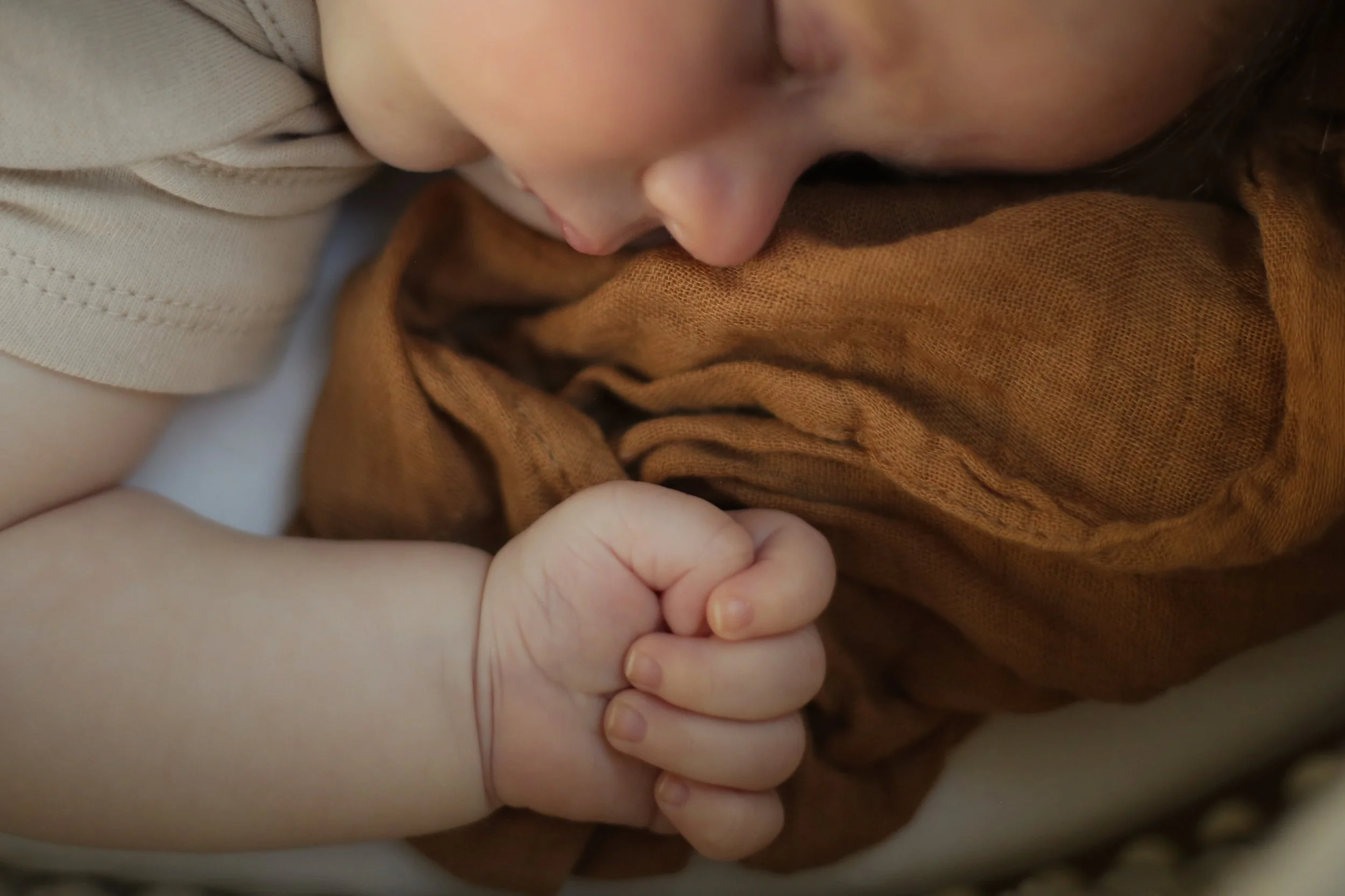 Close-up van een baby die rustig slaapt met haar hoofd tegen een persoon, handen die haar vasthouden, gekleed in een bruin shirt en een beige kledingstuk.