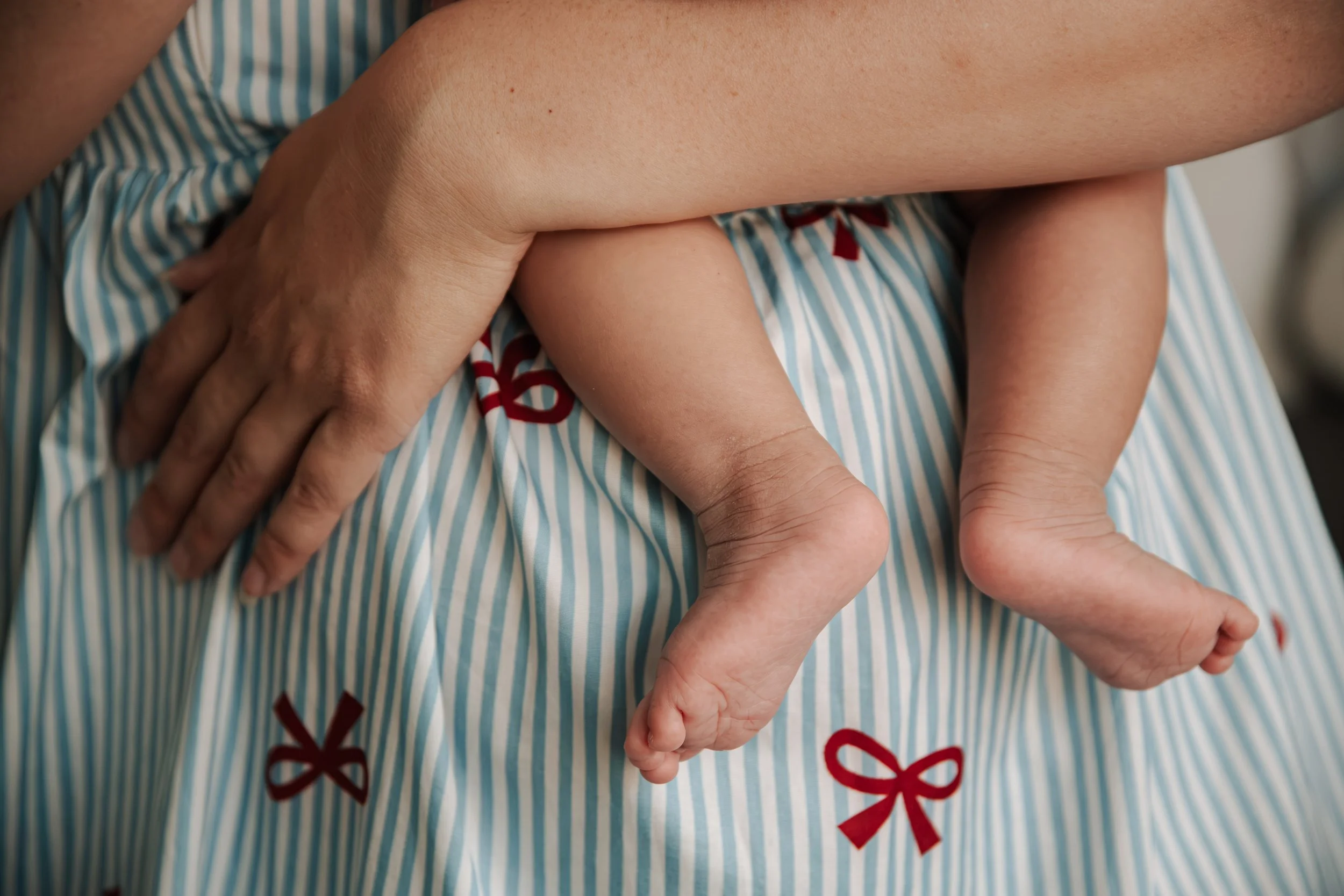 Close-up van een volwassen hand die een kinderbeen vasthoudt, met een baby die ligt op een bed met een blauw-wit gestreepte deken met rode strikjes.