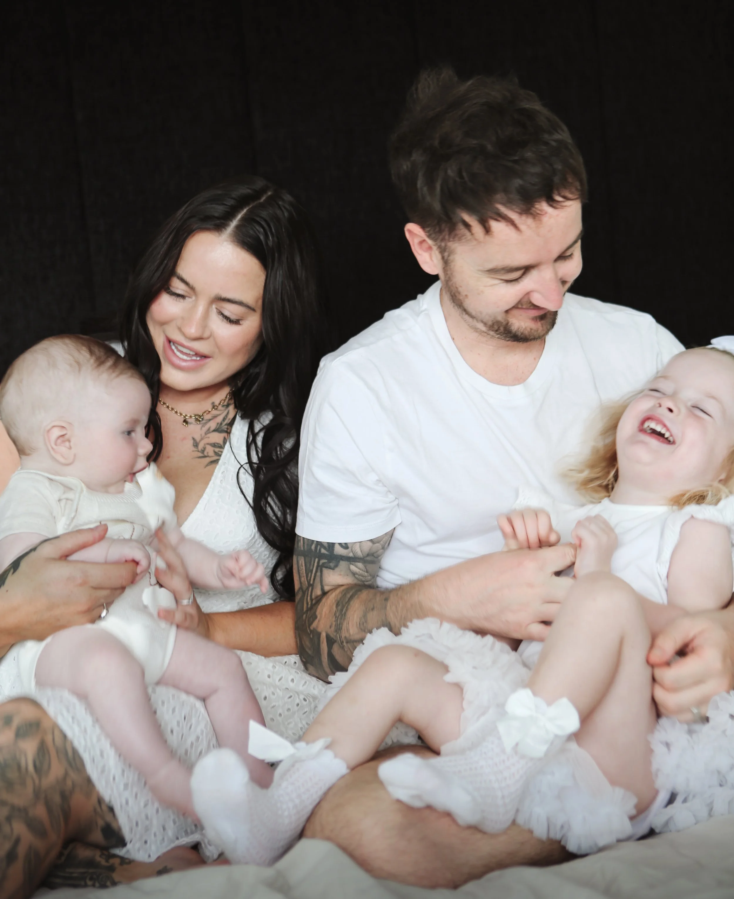 Gezicht van een jonge vrouw met zwart haar, een man met tattoos op de arm, beiden kijken lachend naar baby's in witte kleding en versieringen.