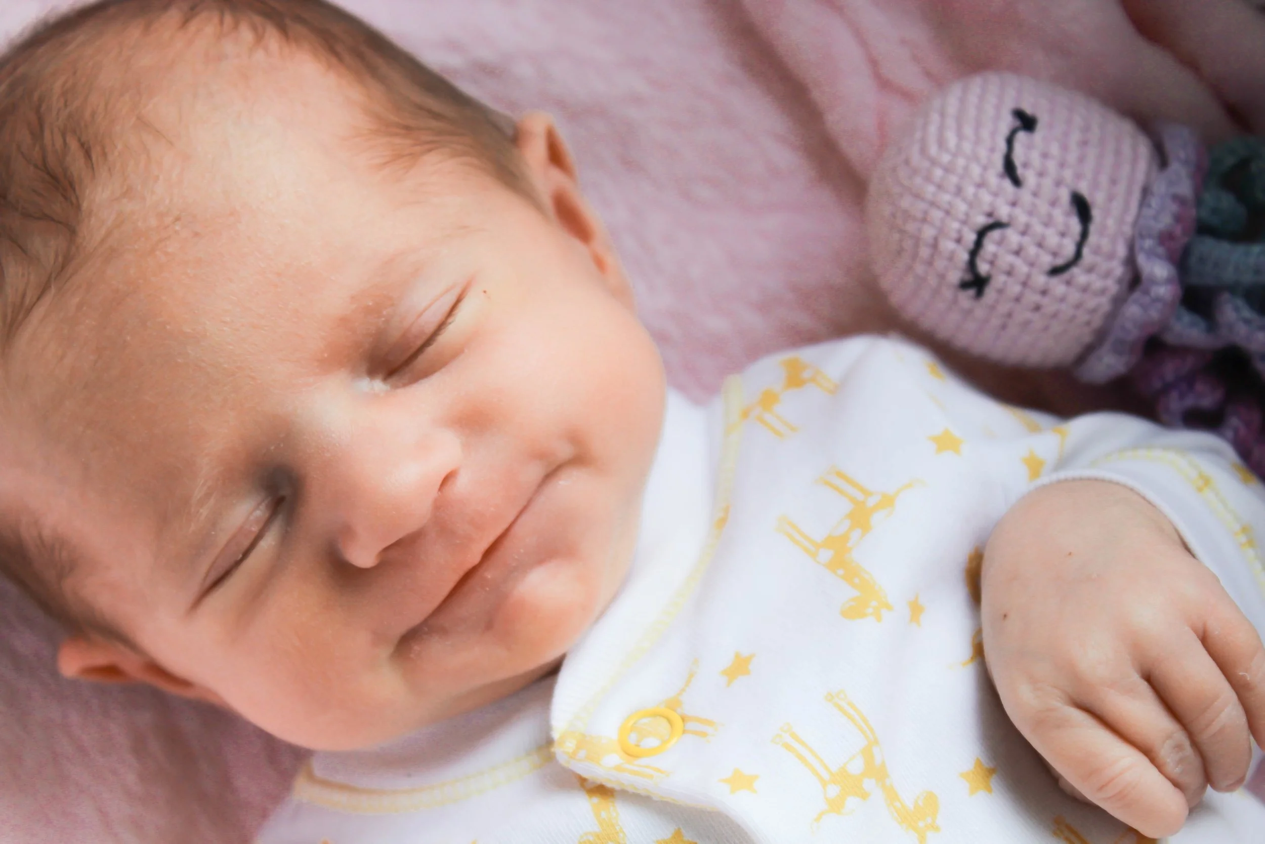 Een slapende baby met lichtbruin haar op een roze deken, gekleed in een witte kleding met gele sterretjes en een kabeltrui met een gezichtje