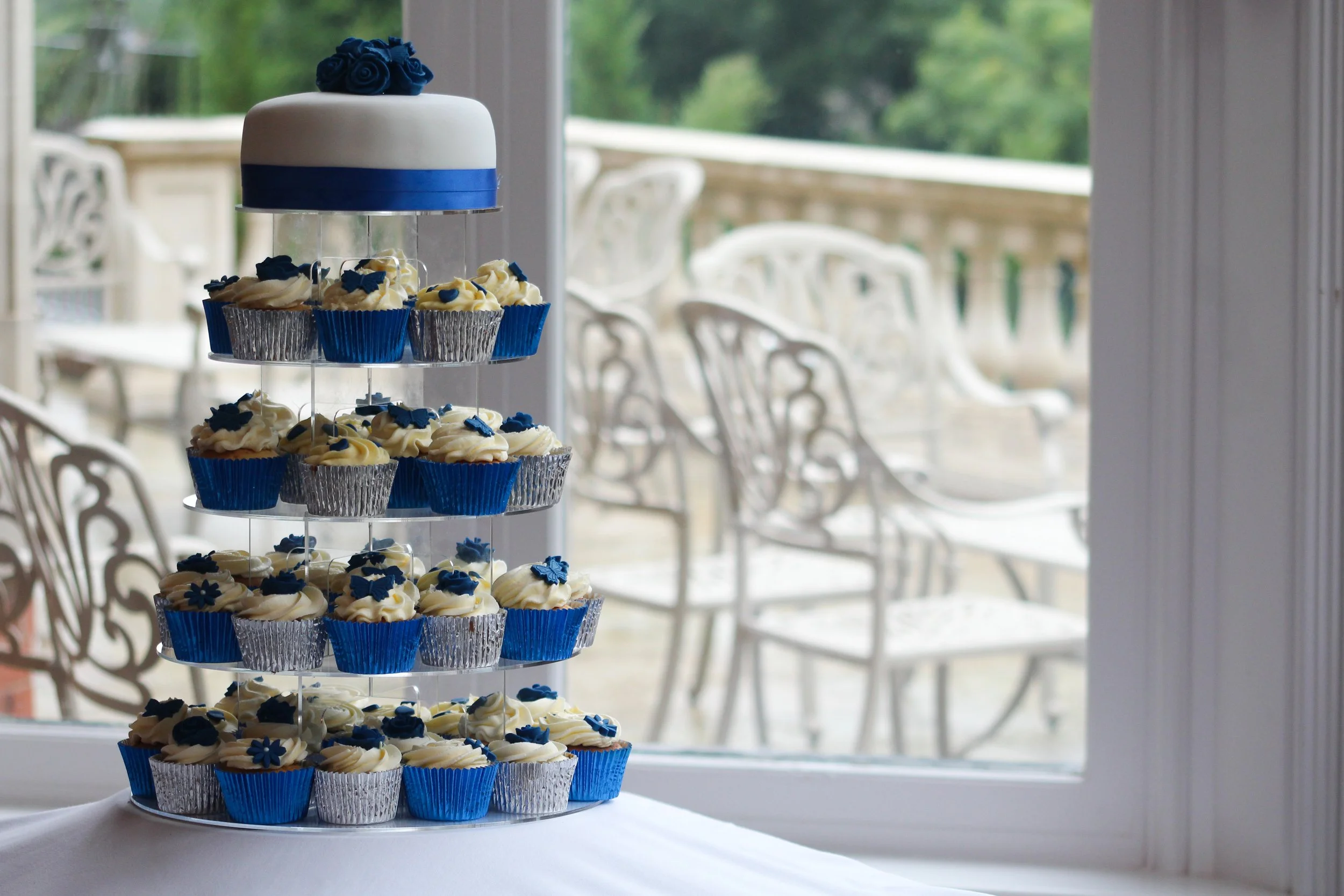 Een taart met blauw en wit ontwerp, met rozen en cupcakes met blauwe en witte decoraties, staat op een tafel bij een raam met uitzicht op een balkon met tuin.