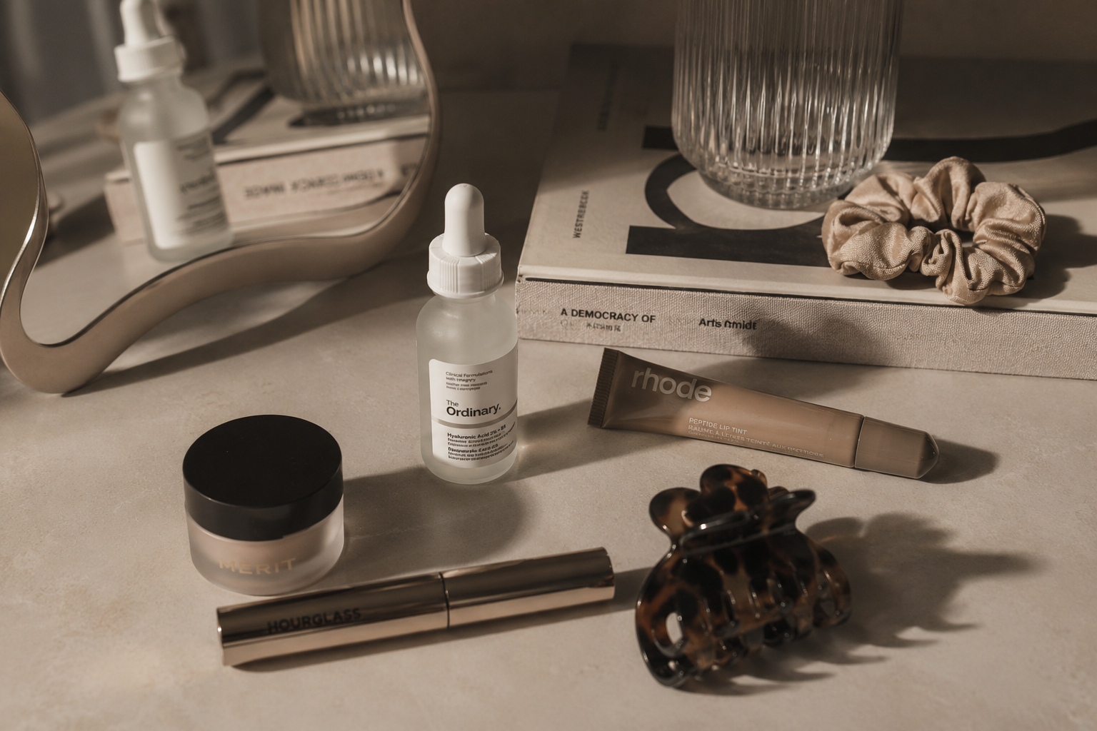 Beauty and skincare products on a beige surface, including a glass bottle of hyaluronic acid, a jar of Merit moisturizer, a tube of Rhodope lip tint, an Hourglass makeup product, a tortoise shell hair clip, a scrunchie, a mirror, a glass vase, and a book with a white cover and black text.