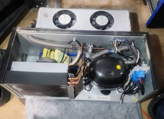 View of an open refrigerator compressor unit with internal components including a black compressor, copper tubing, electrical wiring, and a cooling fan on top.