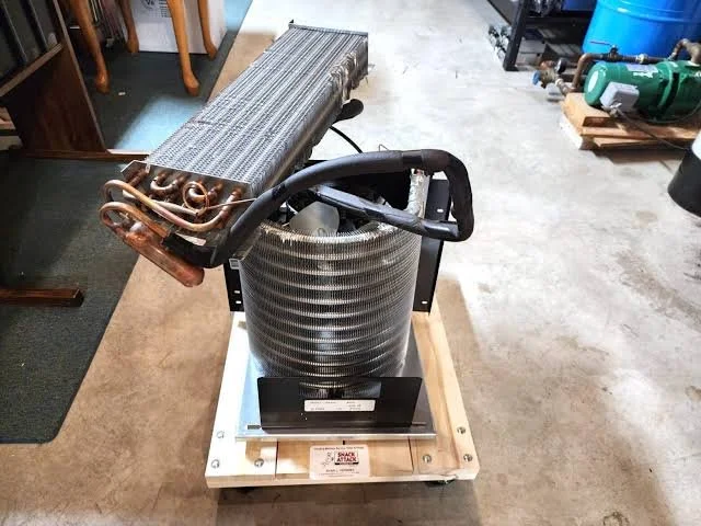 A large cylindrical compressor with a coil of copper pipes and a radiator mounted on top. It is placed on a wooden base on a concrete floor in a workshop or garage.