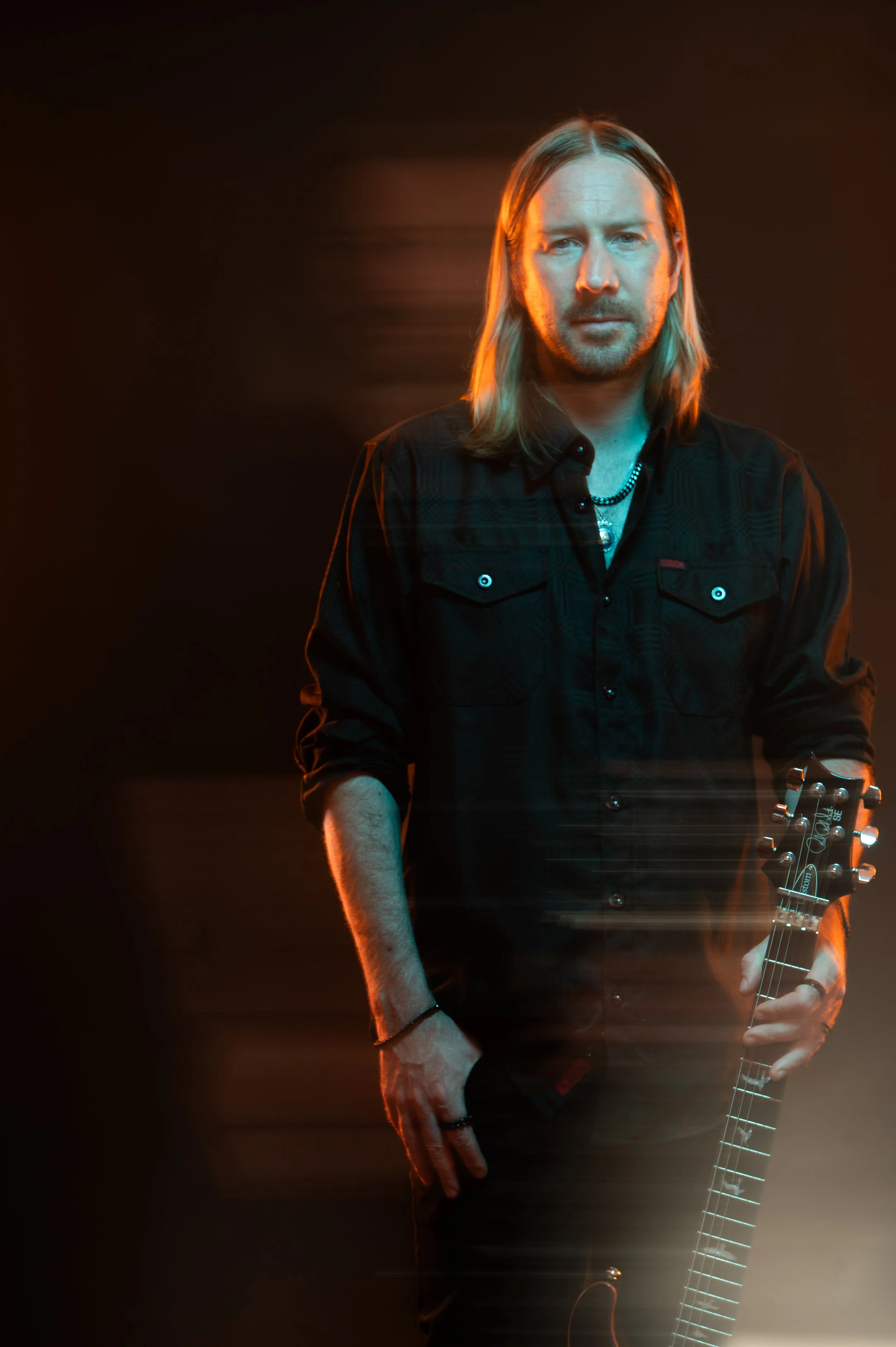 A man with long blonde hair and a beard, wearing a dark plaid shirt and jewelry, stands holding an electric guitar in a dimly lit setting with warm lighting and a dark background.