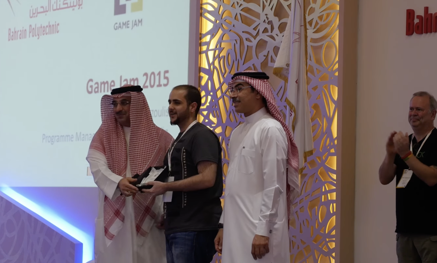 A man in a black T-shirt is receiving an award from a man in traditional Middle Eastern attire with a red and white checkered keffiyeh. Two other men in traditional attire are standing nearby on a stage during an event titled 'Game Jam 2015' at Bahrain Polytechnic.