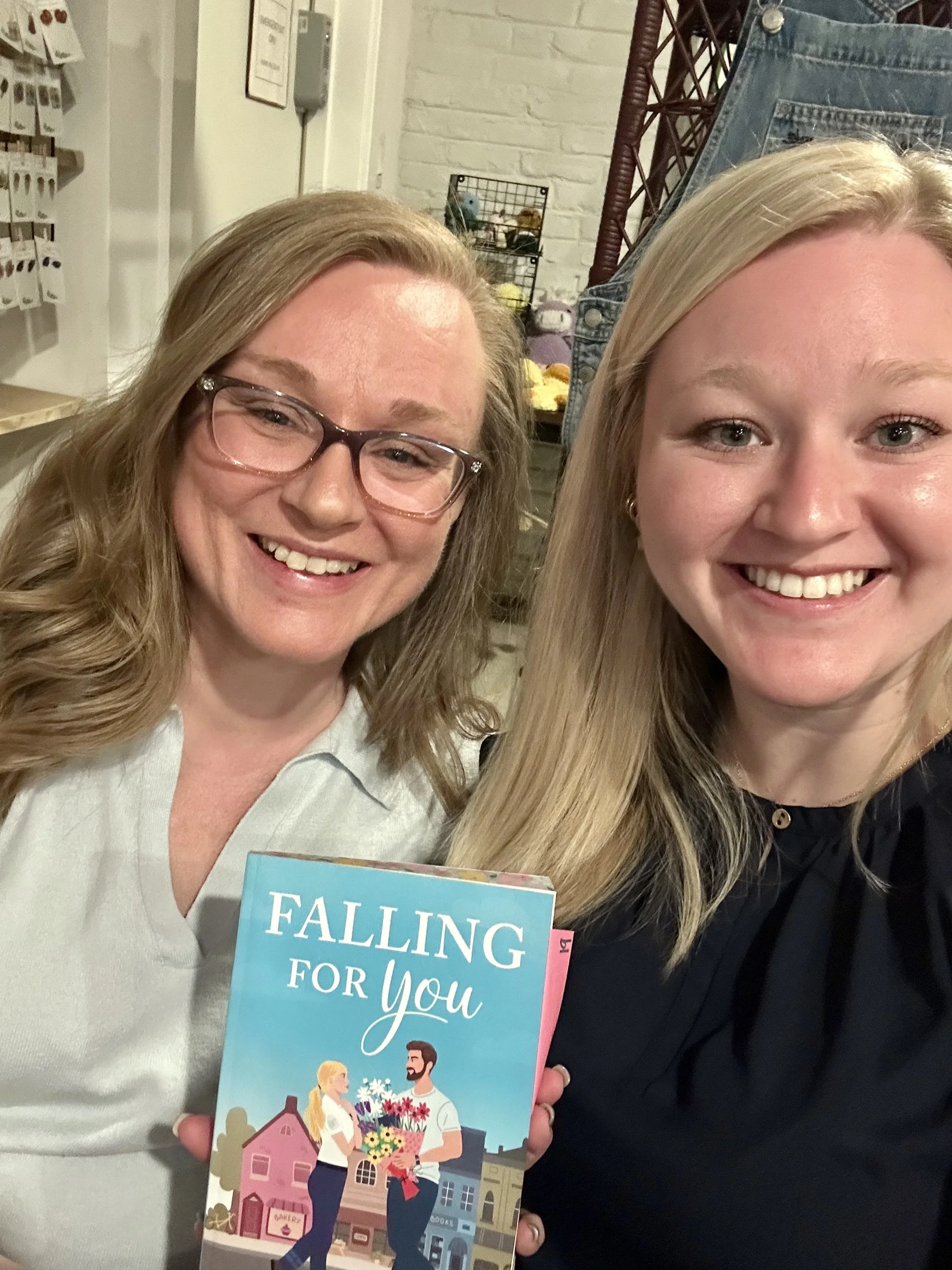 Two women smiling, one holding a colorful book titled four words, 'Falling For You,' in a store with shelves of products and decorative items in the background.