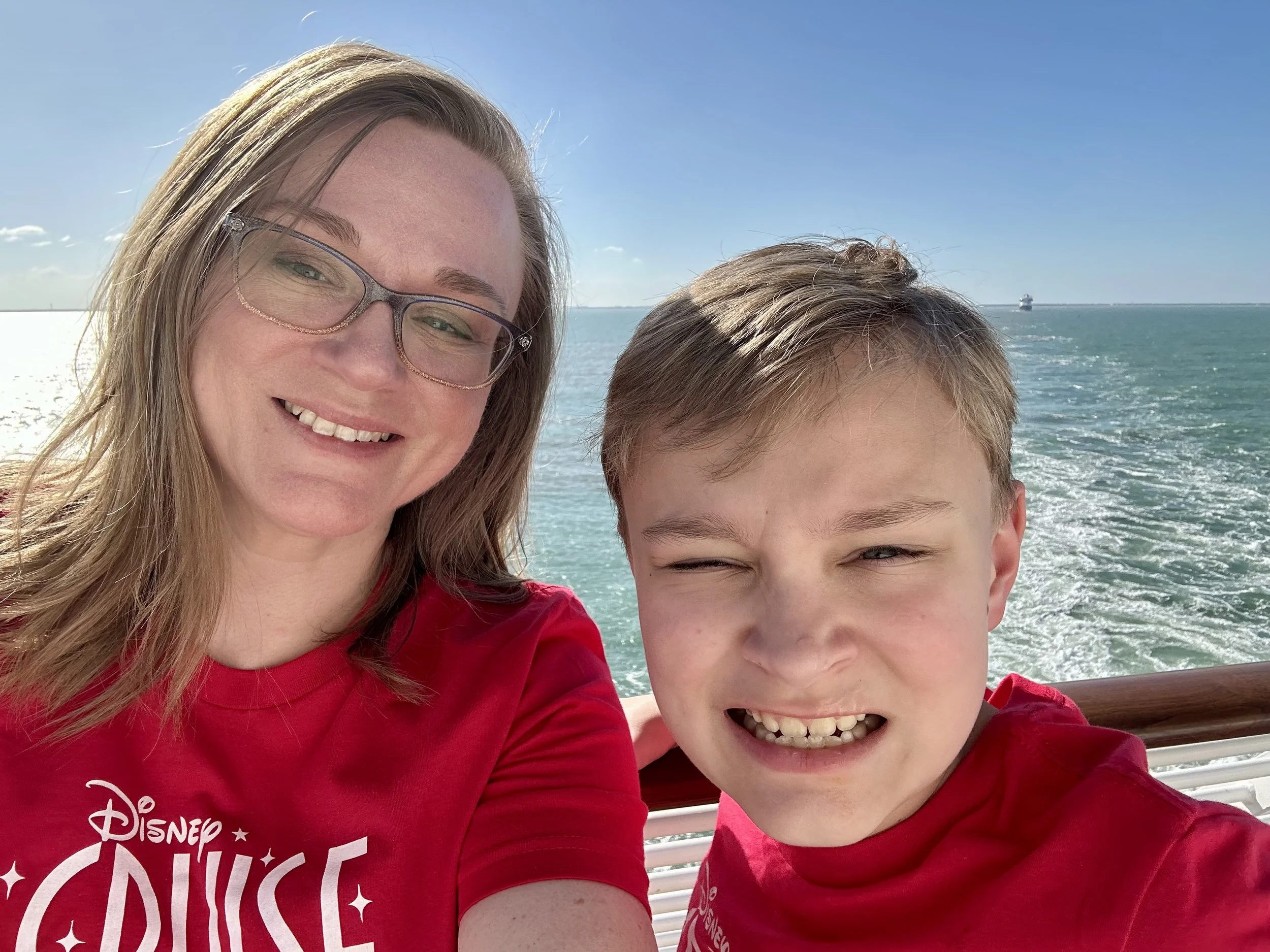 Close-up of a woman with glasses and long hair smiling, and a boy with short hair smiling, on a boat with the ocean and a ship in the background.