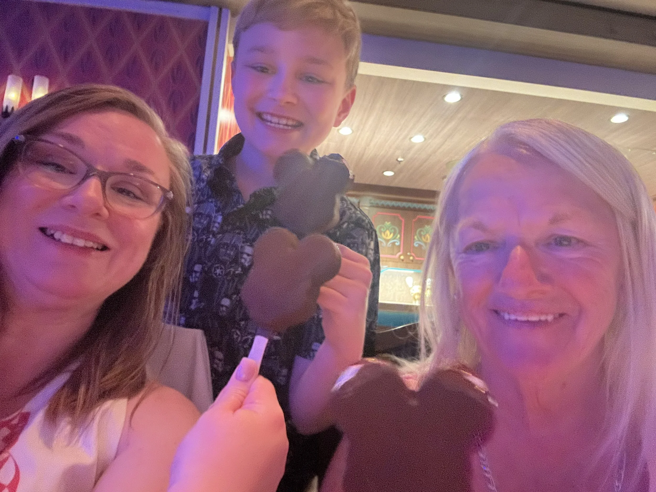 A group of smiling people taking a selfie at an indoor venue with decorative lighting and patterned walls, some giving a thumbs-up and a young boy in the middle holding a heart-shaped item.