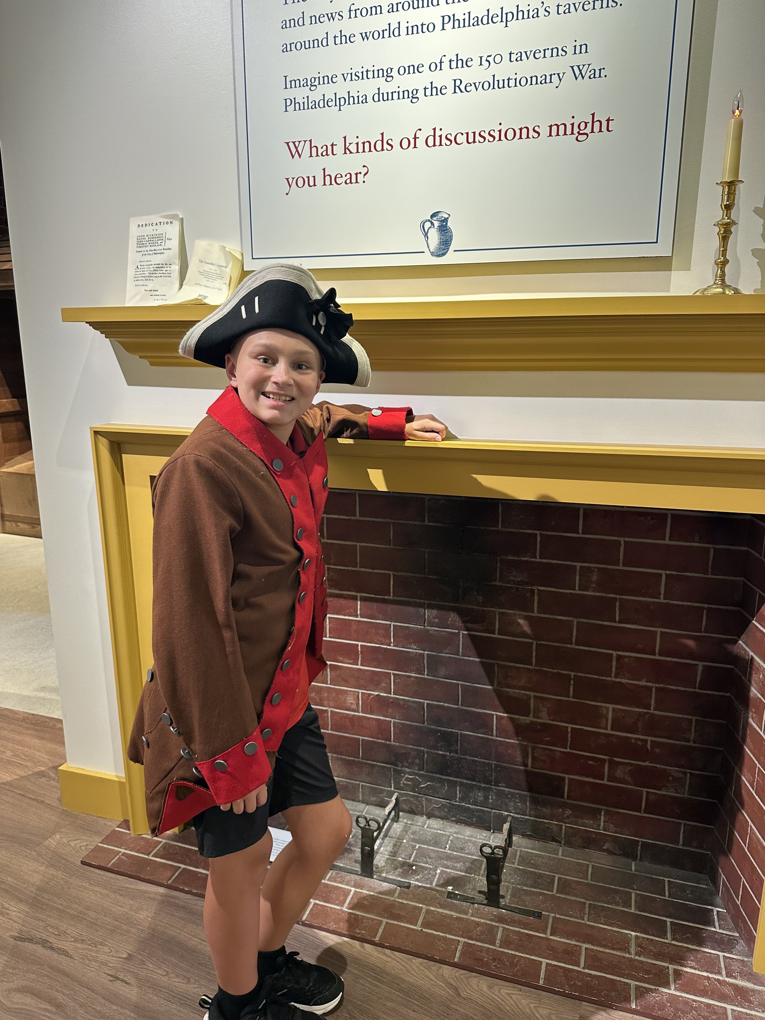 A smiling boy dressed as a Revolutionary War soldier, wearing a tri-corner hat, brown coat with red lining, and black shorts, standing next to a brick fireplace with an empty fire grate and a sign on the wall with text about discussions, in a museum 