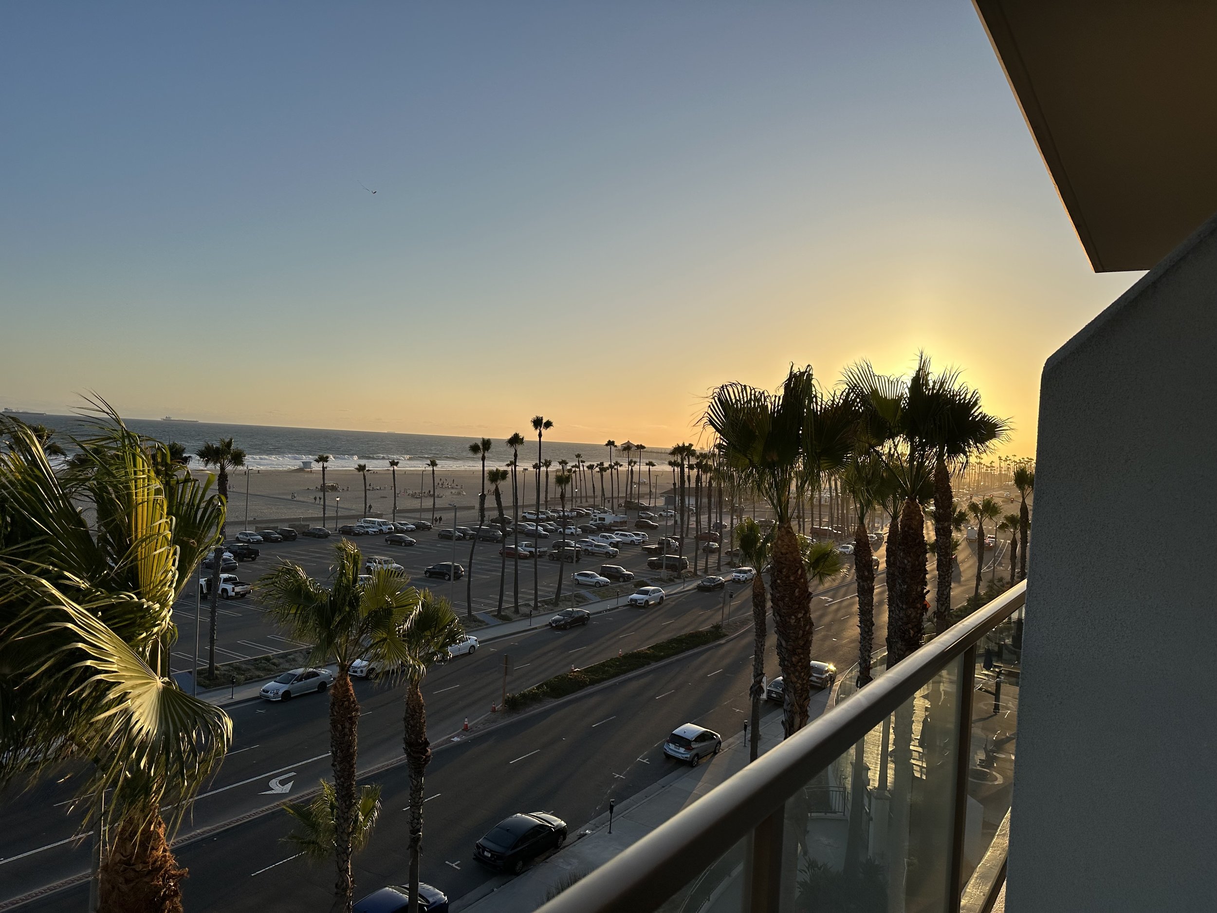 Sunset over a beach with palm trees, a parking lot, and water in the background, taken from a balcony with a metal railing.