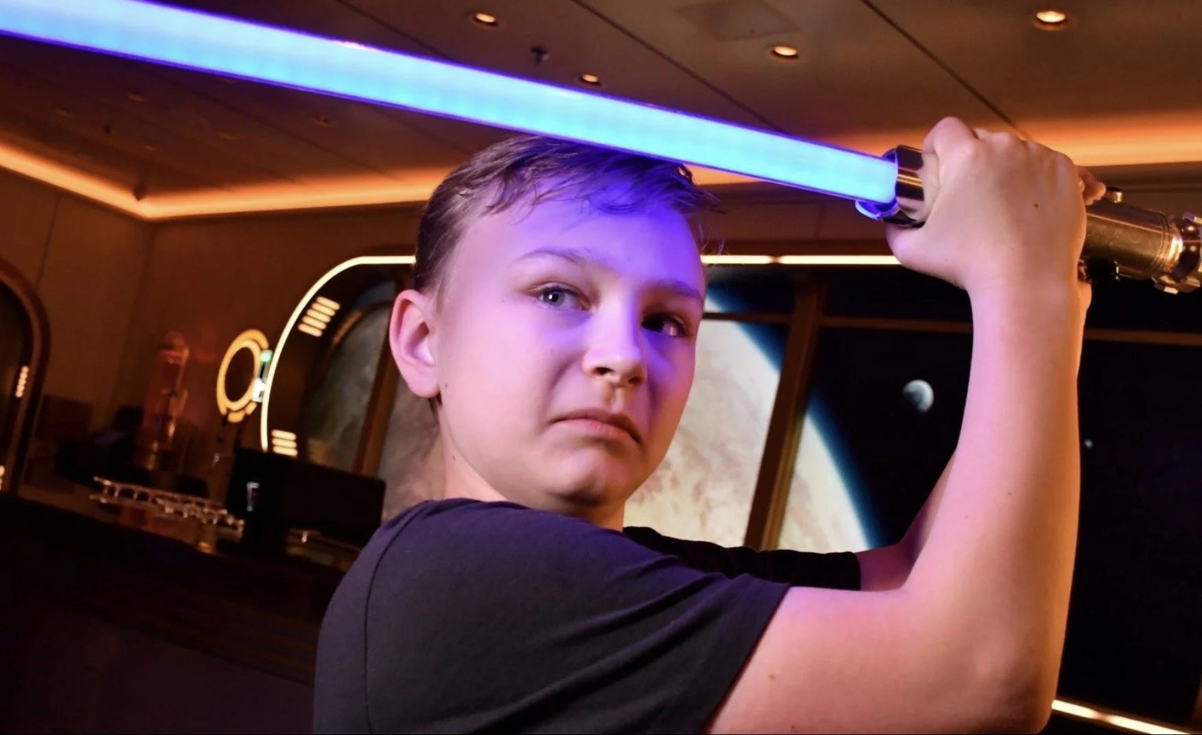 Young person with short hair holding a blue-lit lightsaber in a room decorated with space-themed artwork.