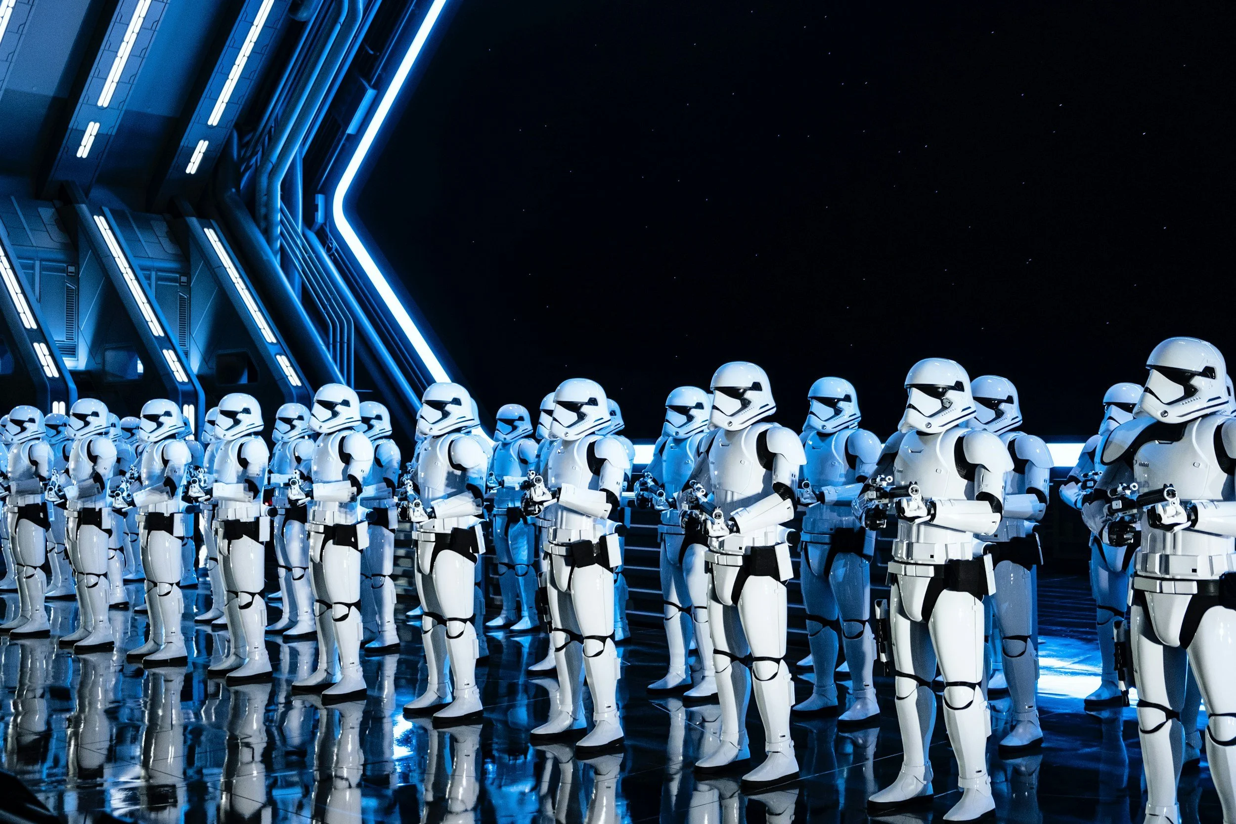 Line of stormtroopers in white armor on a reflective surface at night, with a futuristic building illuminated by blue lights next to a starry sky.