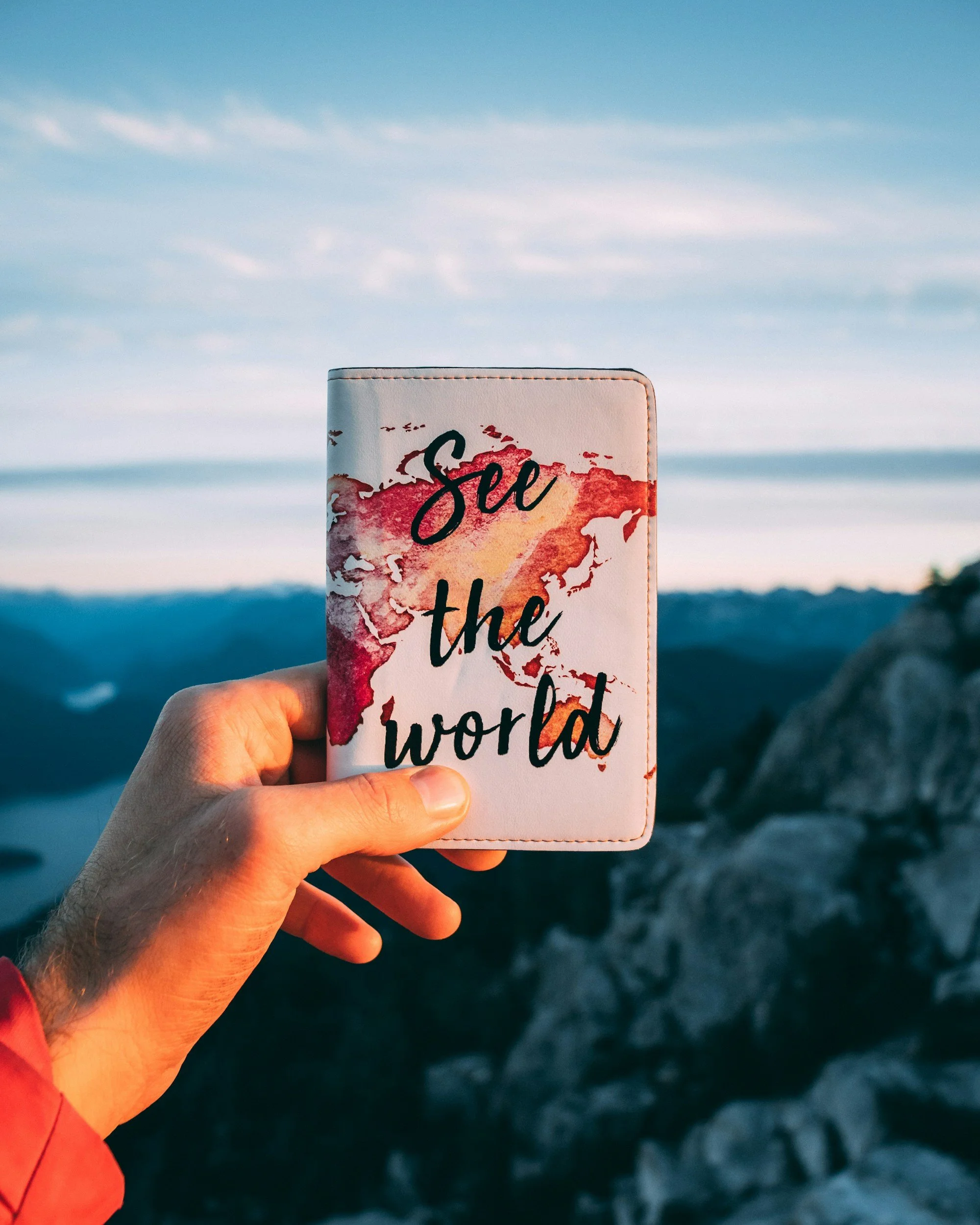Hand holding a world map passport holder with the phrase 'See the world' written on it, against a backdrop of a sky and distant mountains.