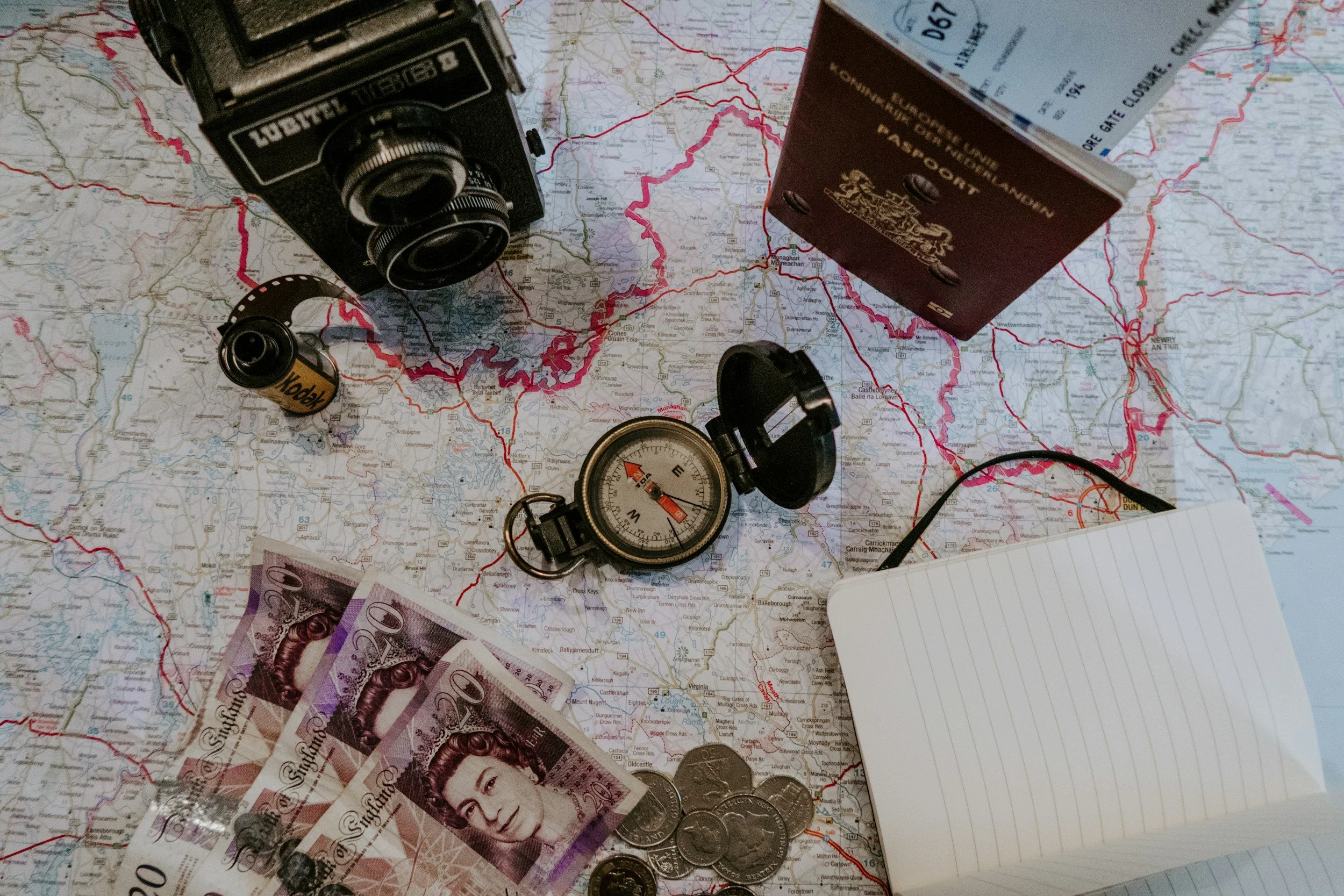 A vintage camera, a bottle of whiskey, a compass, several 20-pound banknotes, and coins placed on a map with red and black routes.