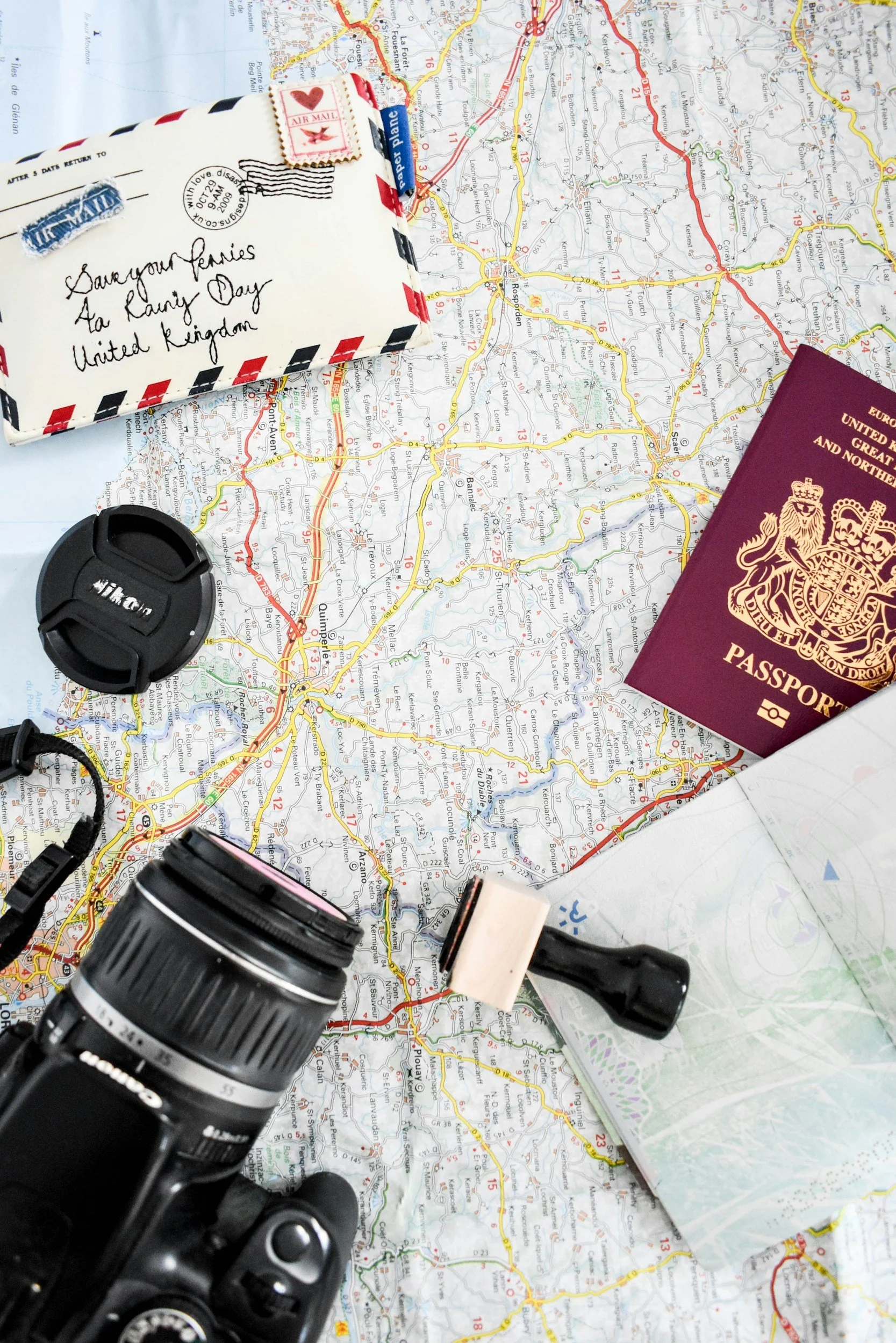 Travel essentials including a map of France, a camera, a camera lens, a backpack, a passport, and an envelope with a letter and a small package with a postage stamp, placed on top of the map.