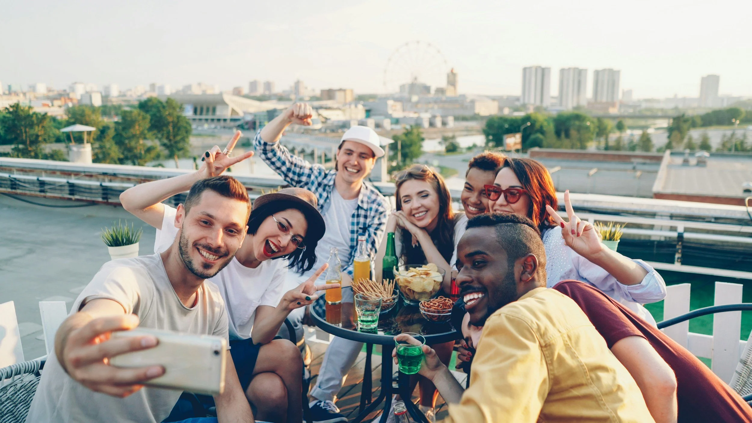 Group of friends smiling and taking selfies at a rooftop party with city skyline in the background.