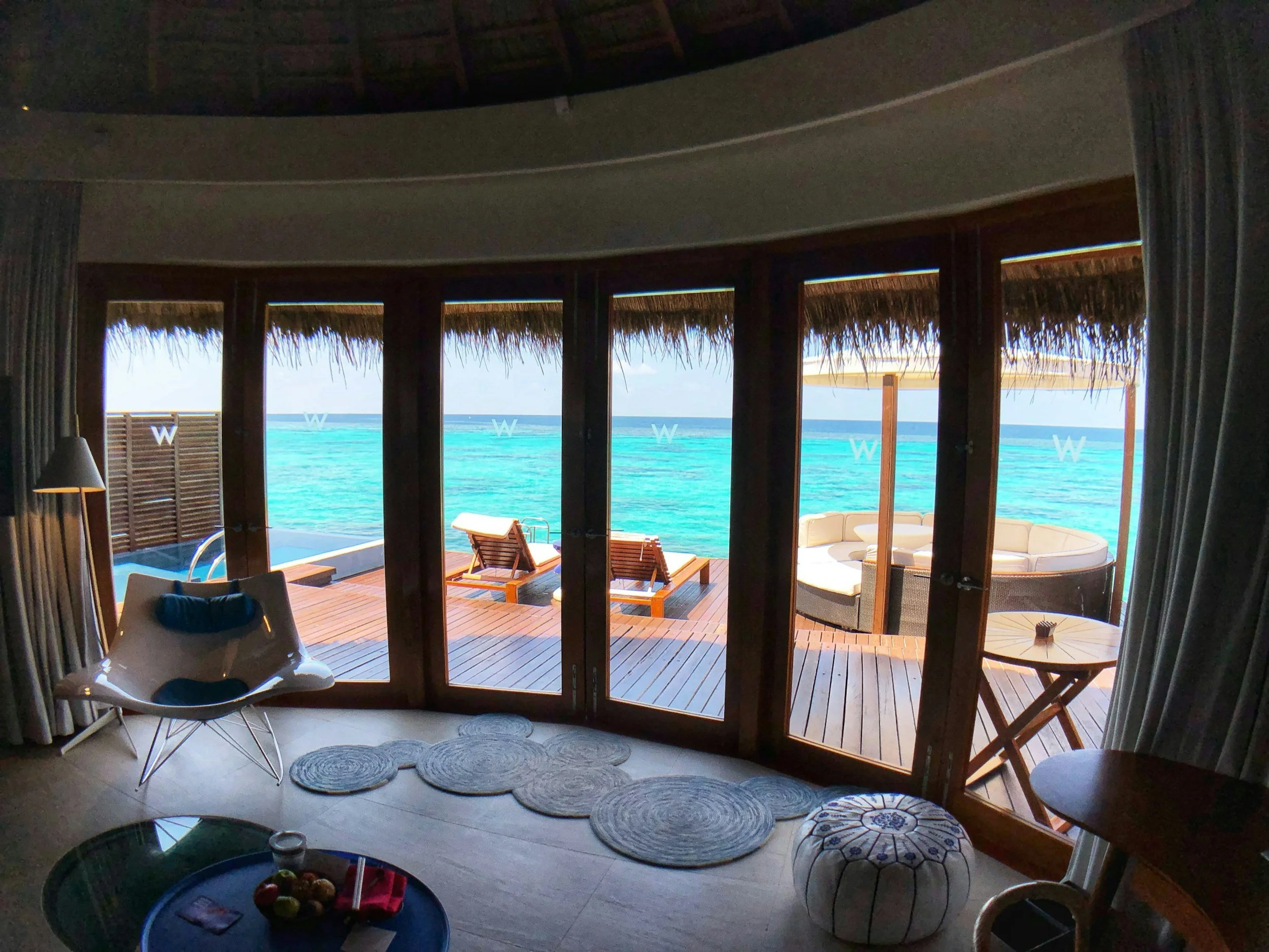 Interior view of a room with sliding glass doors opening to a tropical beach with turquoise water. The room has a white chair in the foreground, a small table with fruit, and outdoor furniture including lounge chairs and a circular sofa with cushions on a wooden deck.